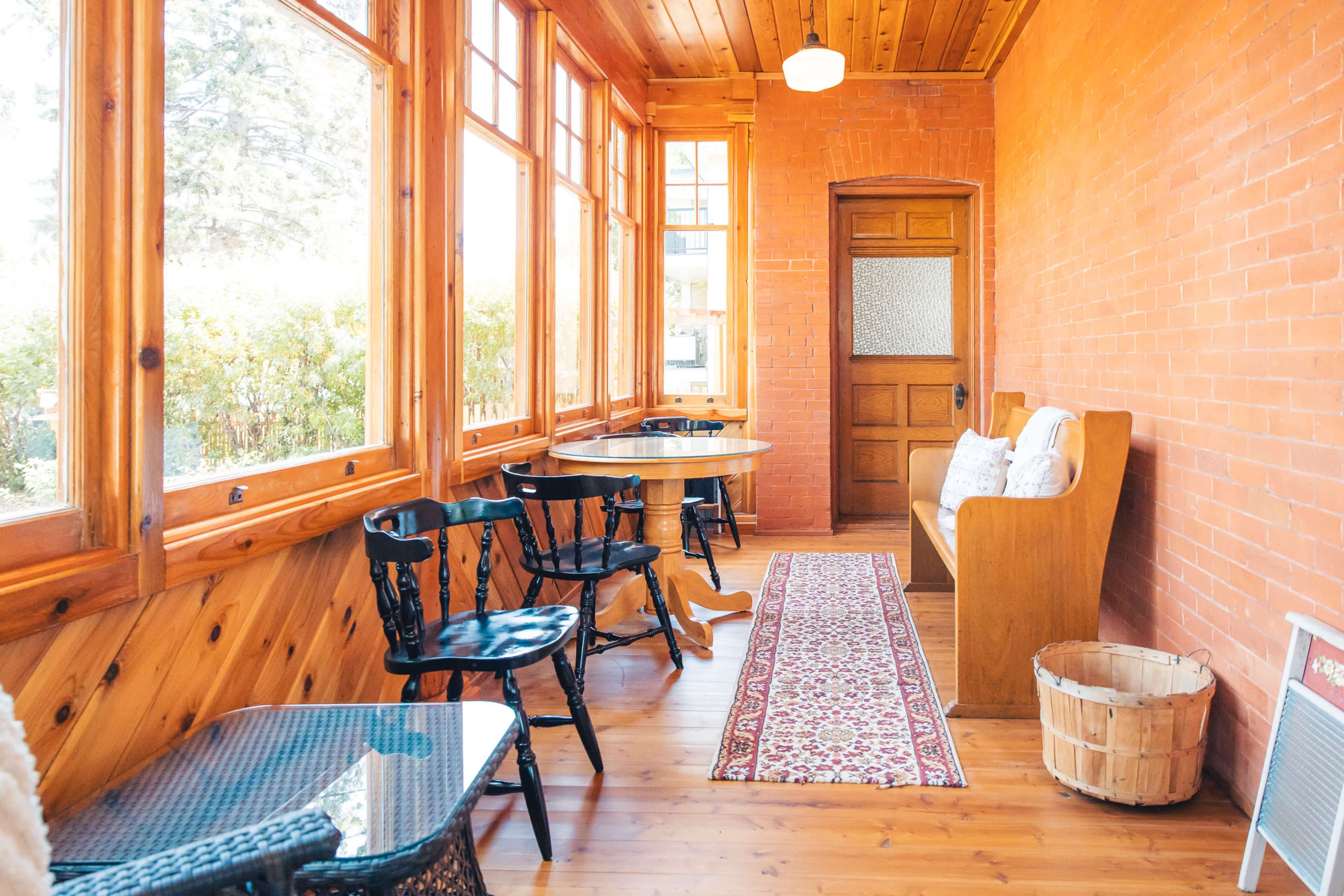 The image shows a sunlit sunroom with brick walls, wooden floors, and a combination of seating arrangements, including chairs and a bench.