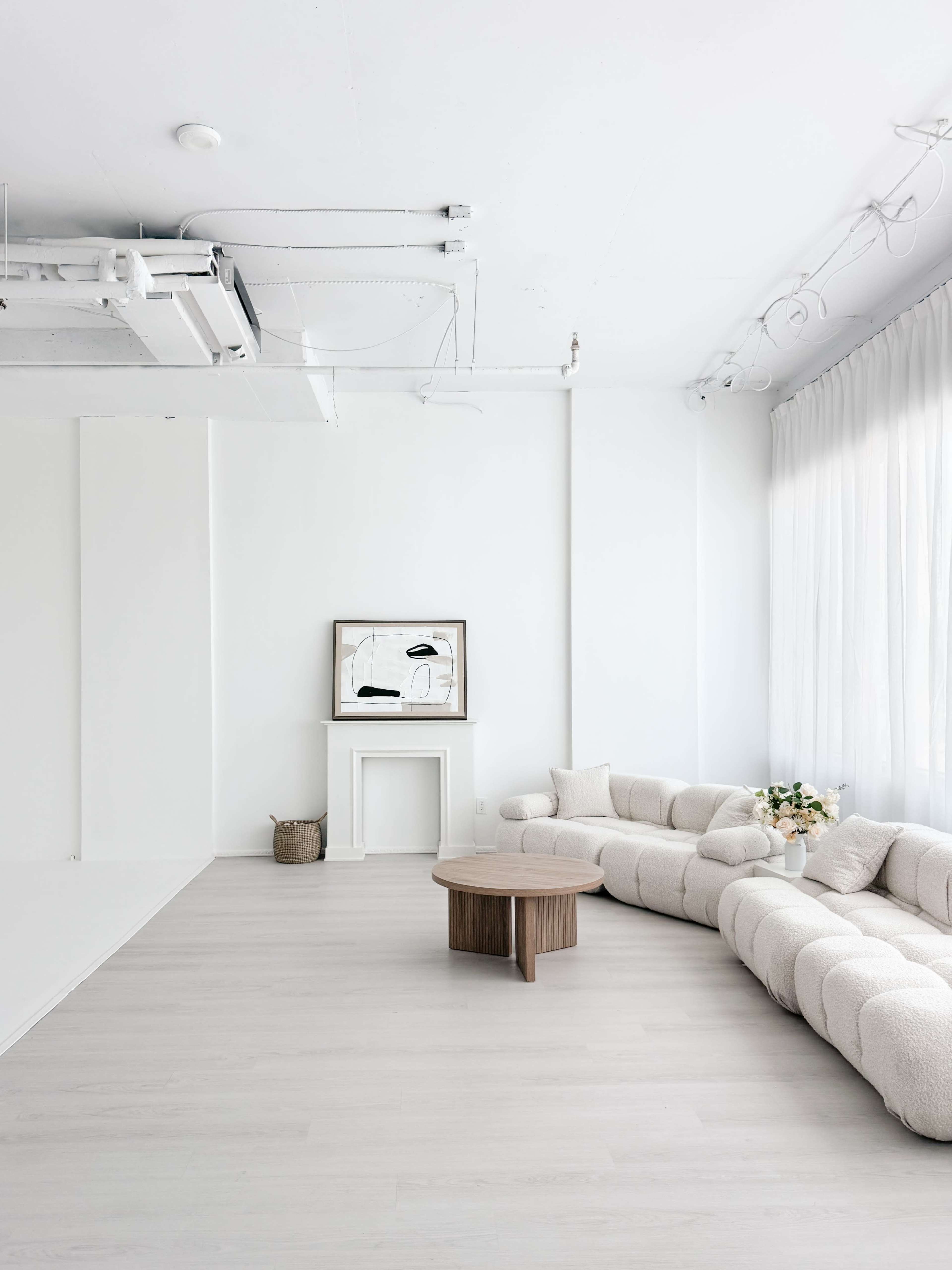 The image shows a minimalistic room with a large, light-colored sectional sofa, a round wooden coffee table, and a framed artwork on the wall, along with natural light coming through sheer curtains.