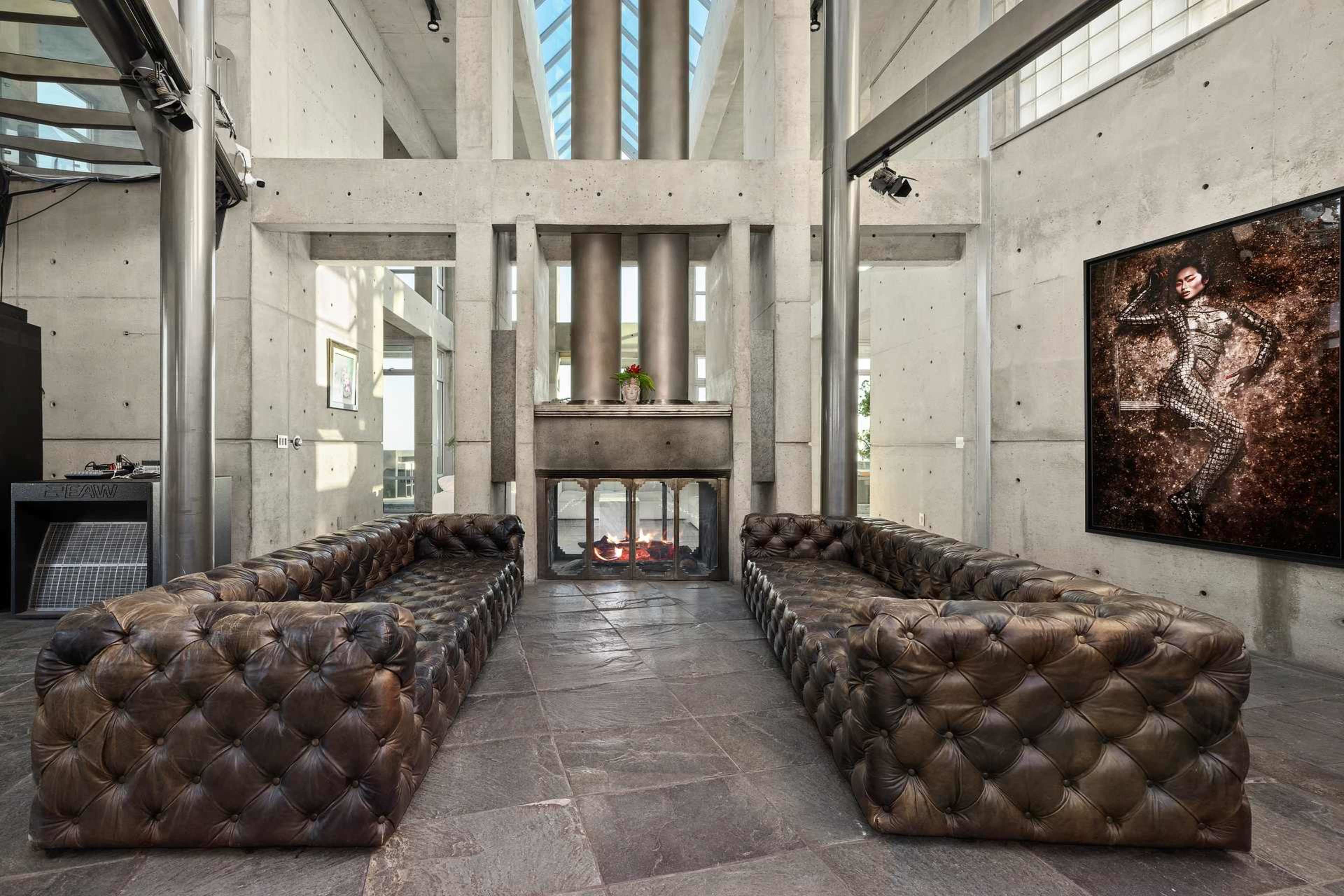 The image shows a modern interior space with two tufted leather sofas facing a fireplace, surrounded by concrete walls and large windows.