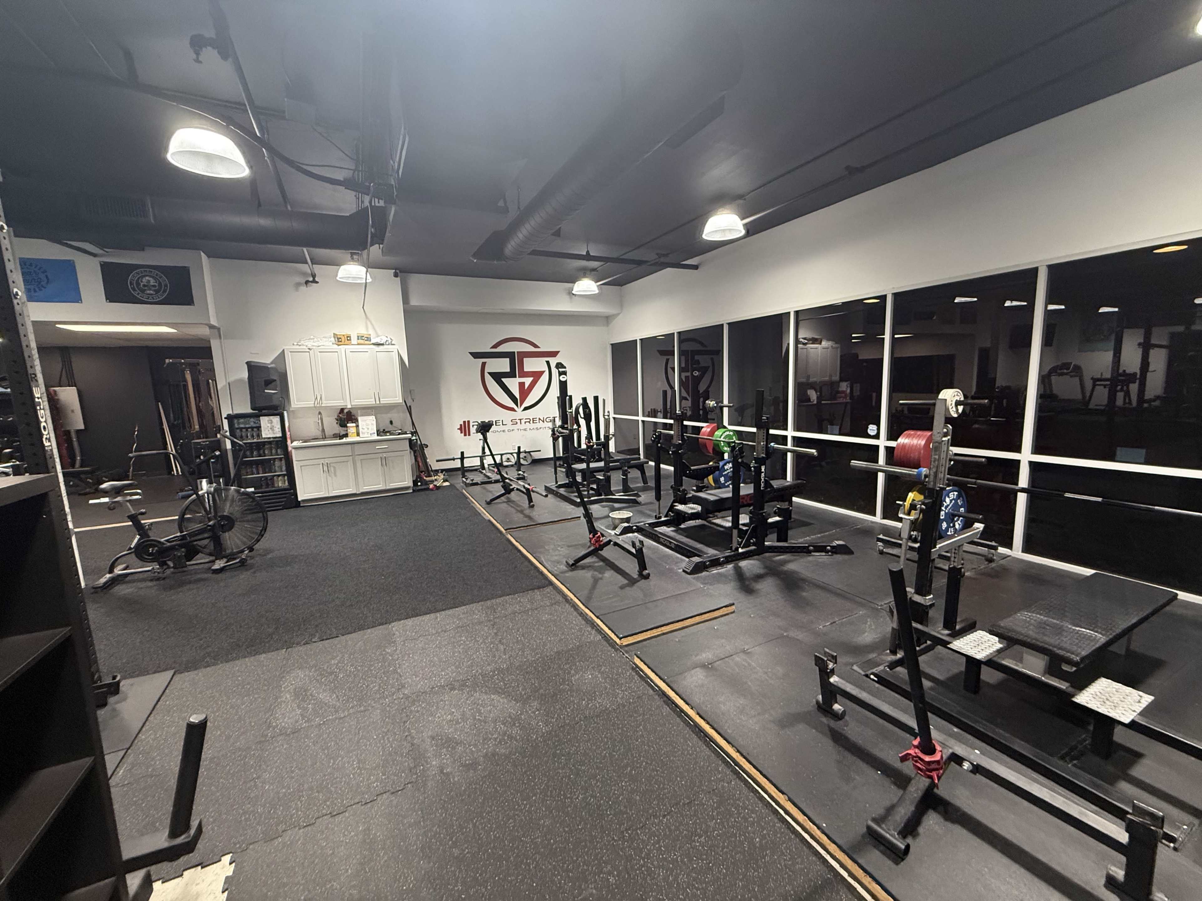 The image shows a well-equipped gym interior featuring weightlifting racks, benches, and various fitness equipment arranged on a rubberized floor.