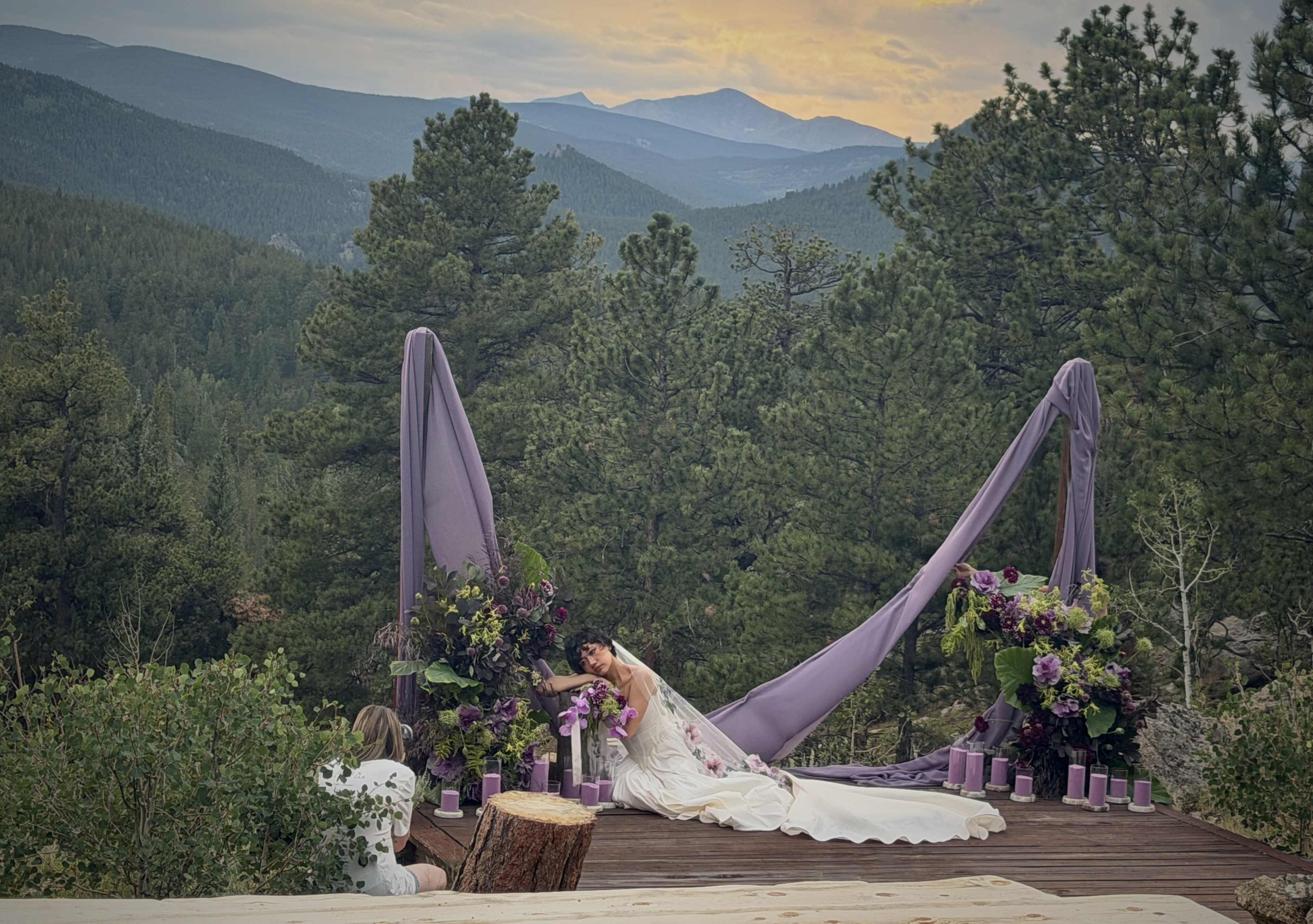A woman in a white dress poses near a decorative backdrop of purple drapes and flowers on a wooden platform overlooking a mountainous landscape at sunset.