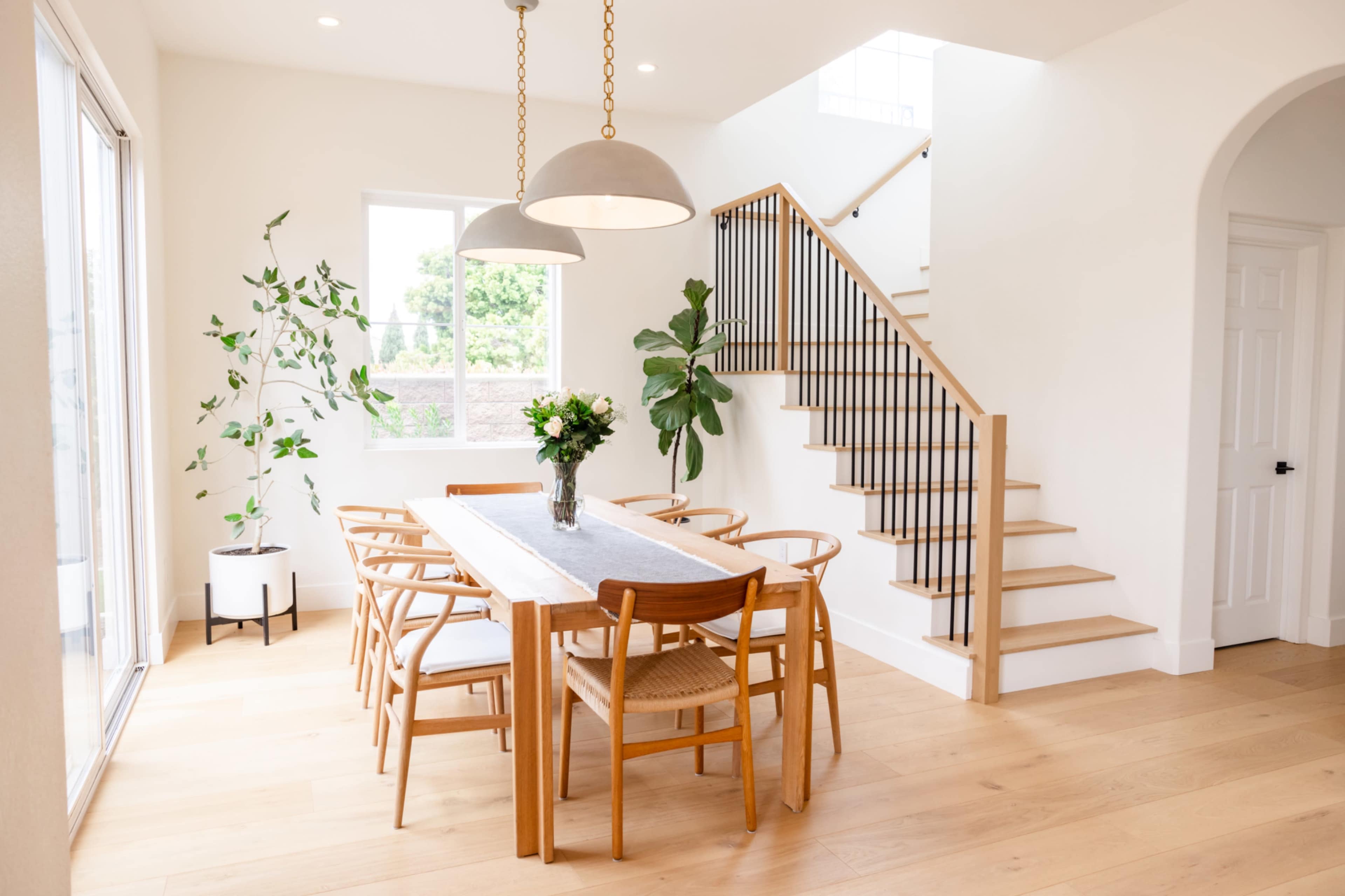 A spacious dining area features a wooden table surrounded by chairs, a vase of flowers in the center, and a staircase leading to an upper level.