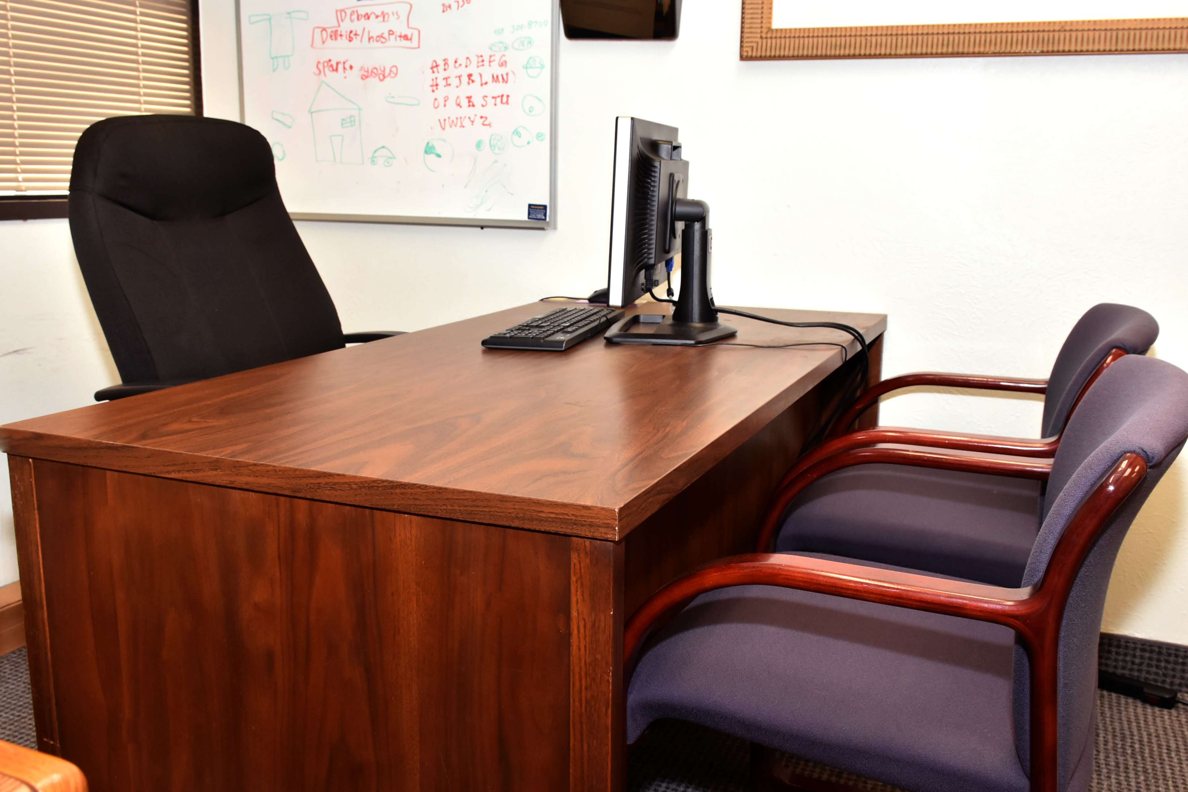 A wooden desk with a computer and keyboard in an office, accompanied by an ergonomic chair and two purple upholstered chairs.