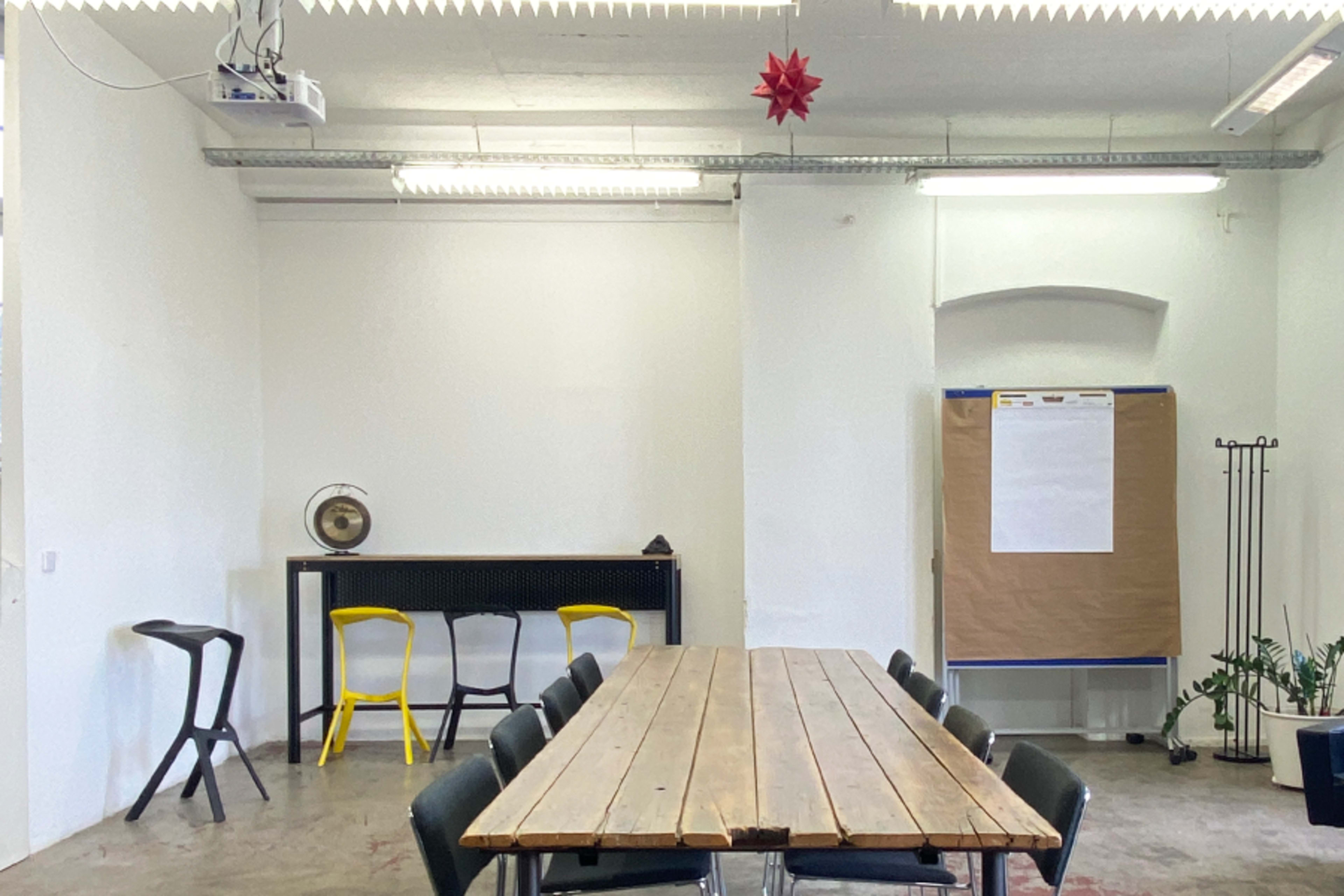 A spacious meeting room features a long wooden table surrounded by black chairs, a small bar with yellow stools, and a presentation area with a board on the wall.