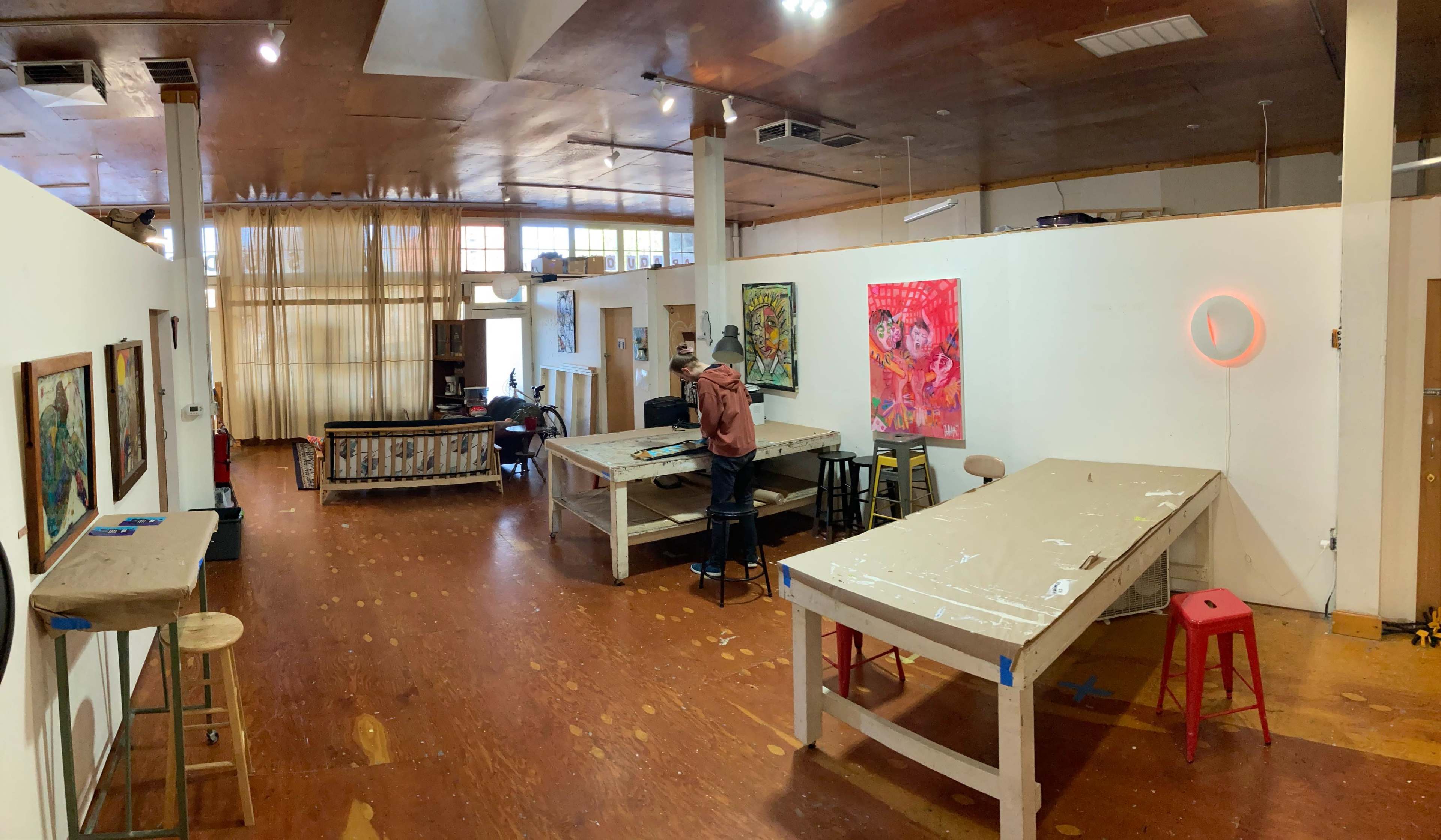 The image shows an artist studio with several tables, artwork on the walls, and individuals working in separate areas.