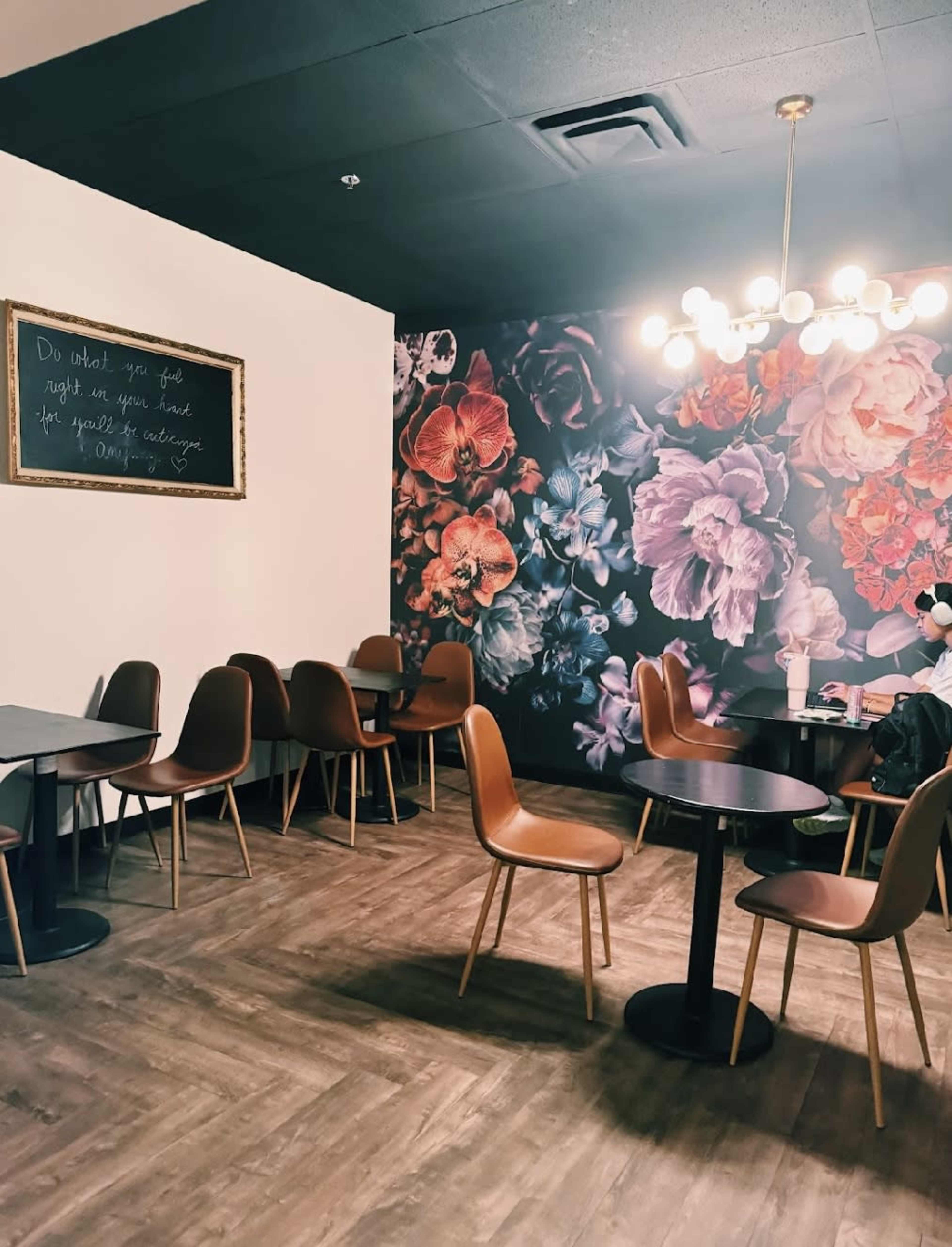 The image shows a cafe interior with several brown chairs and round tables, featuring a floral mural on one wall and a chalkboard with text on another.