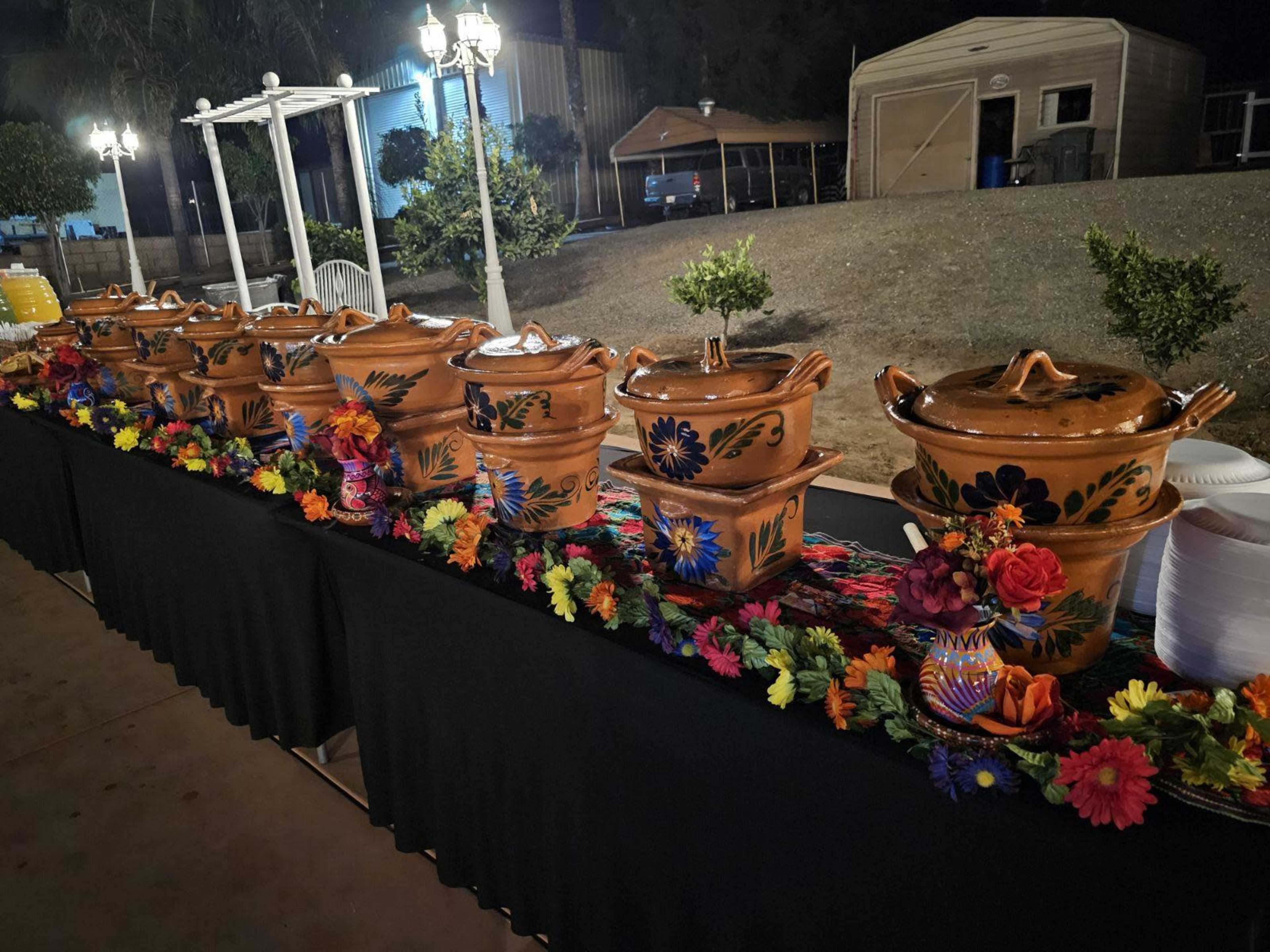 A long table is set with colorful clay serving pots and decorative flowers for a buffet-style meal at night.