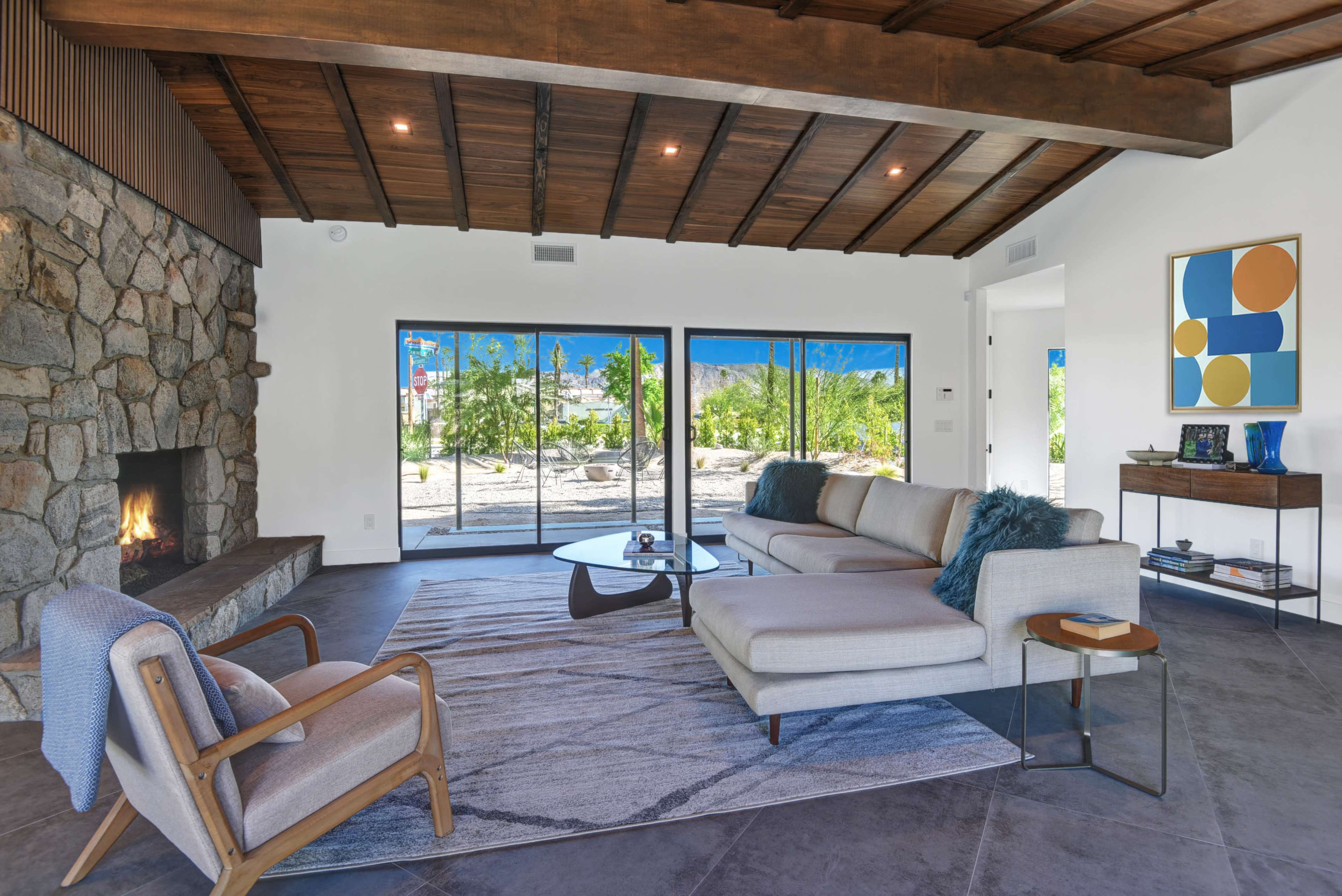 A contemporary living room features a stone fireplace, large glass doors opening to a landscaped view, and modern furniture arranged around a circular coffee table.