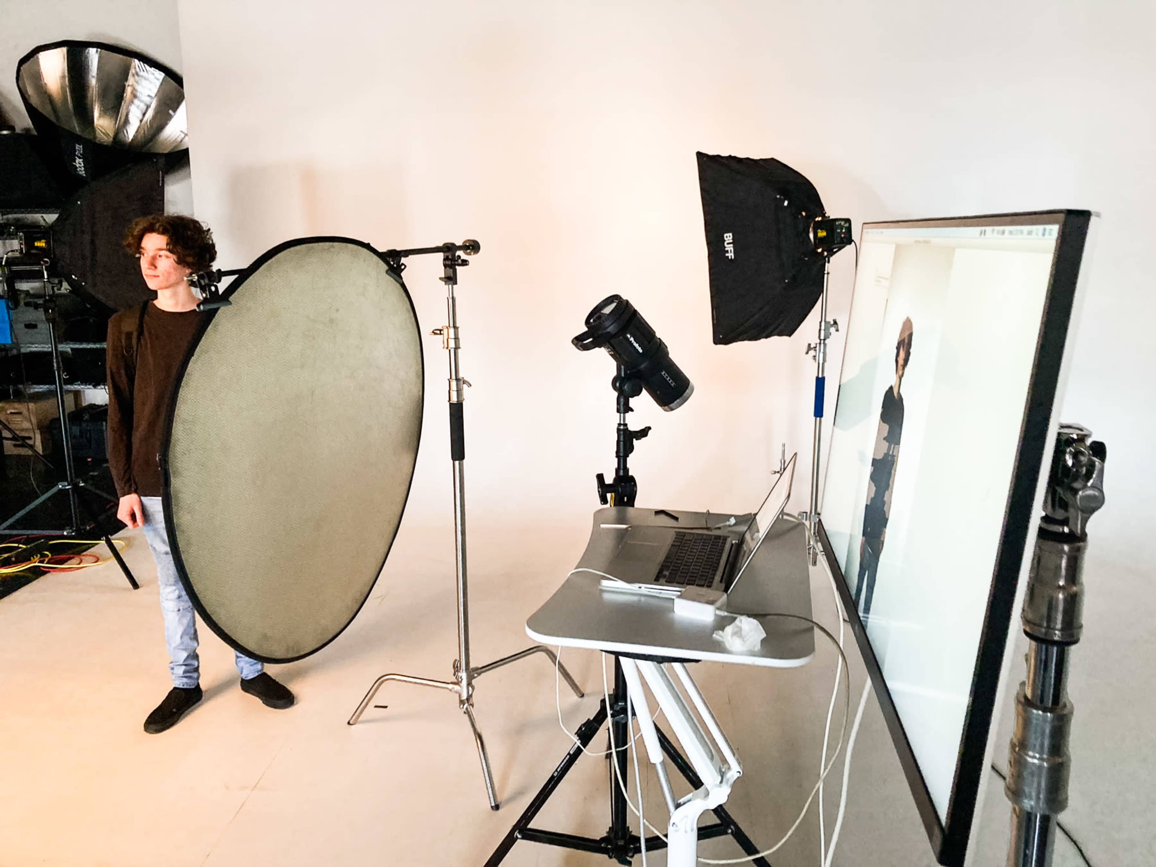 A male model stands beside a large reflector in a photography studio equipped with lighting and a computer displaying a live image.