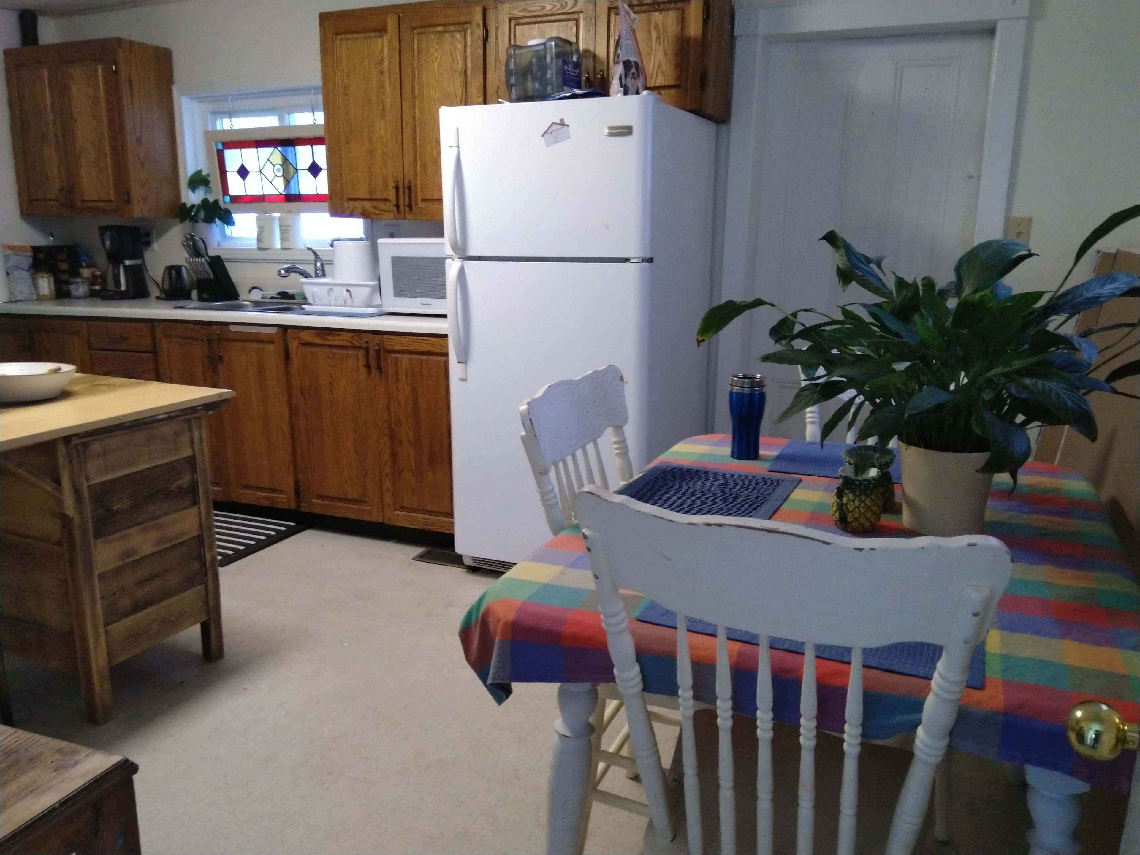 A cozy kitchen features wooden cabinets, a white refrigerator, a dining table with a colorful checkered cloth, and a potted plant.