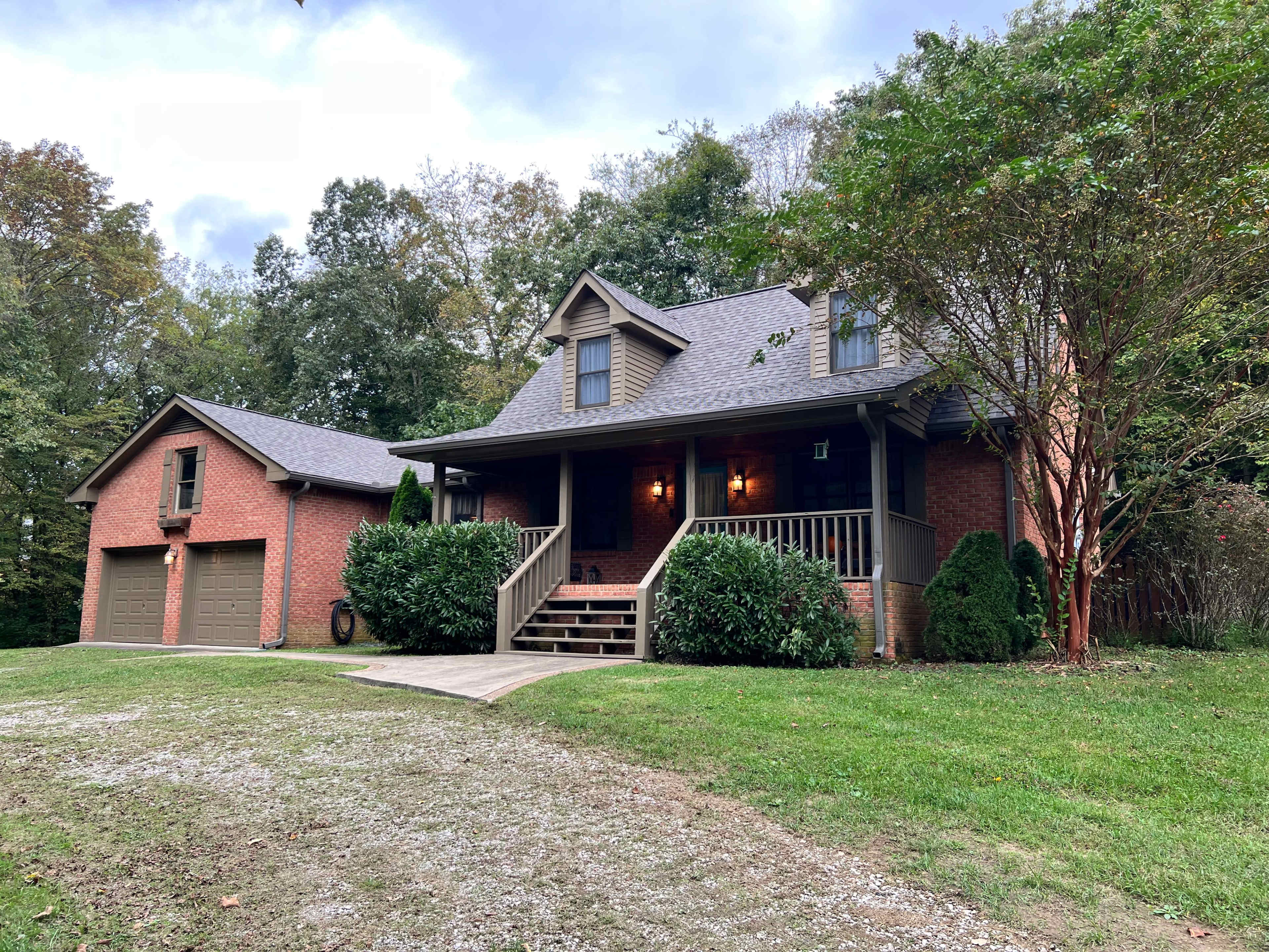 A brick house with a front porch and a garage is surrounded by trees and grass.