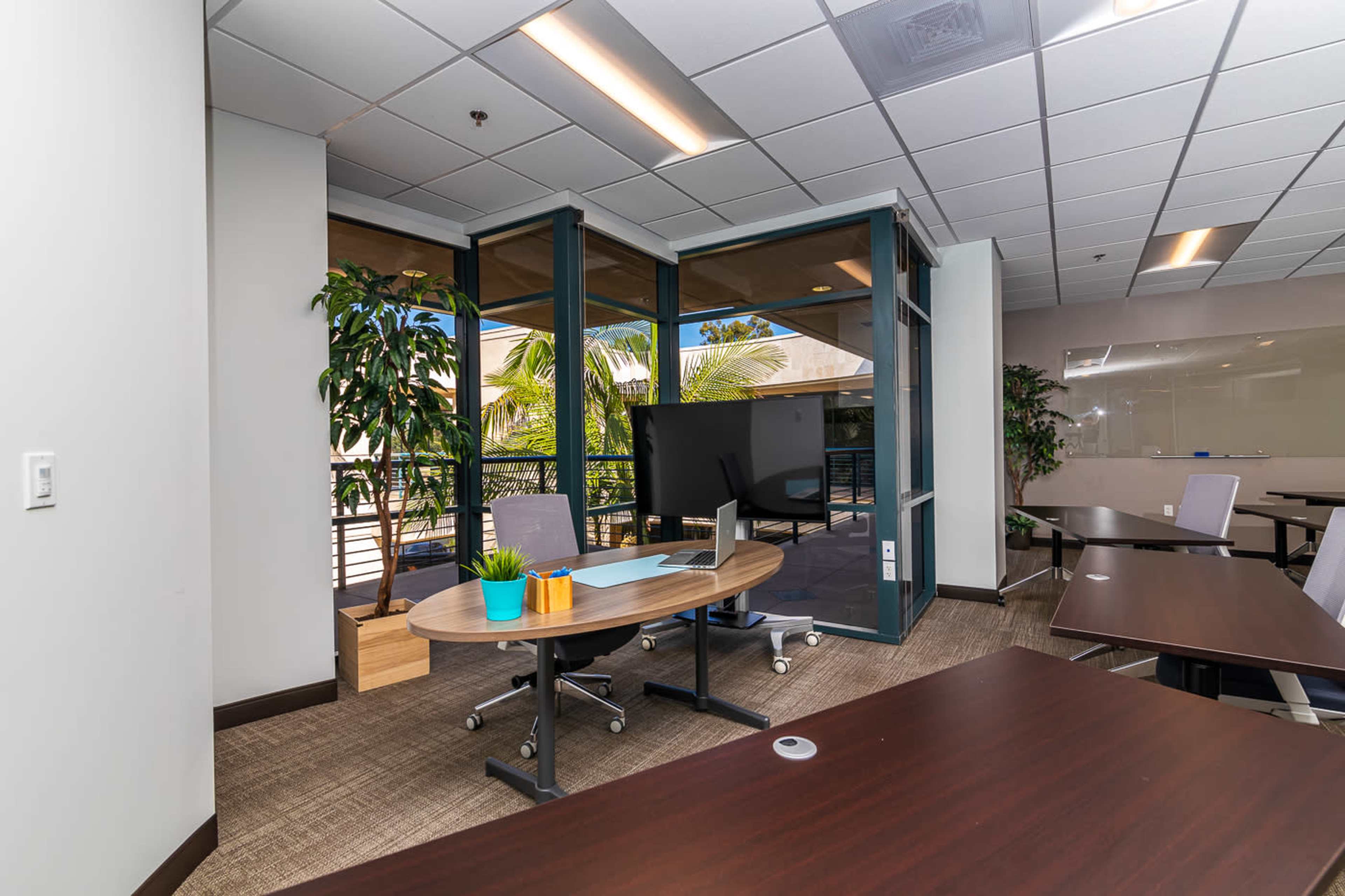 A modern office space with glass doors, a large screen on a stand, and multiple tables arranged for meetings, surrounded by greenery.