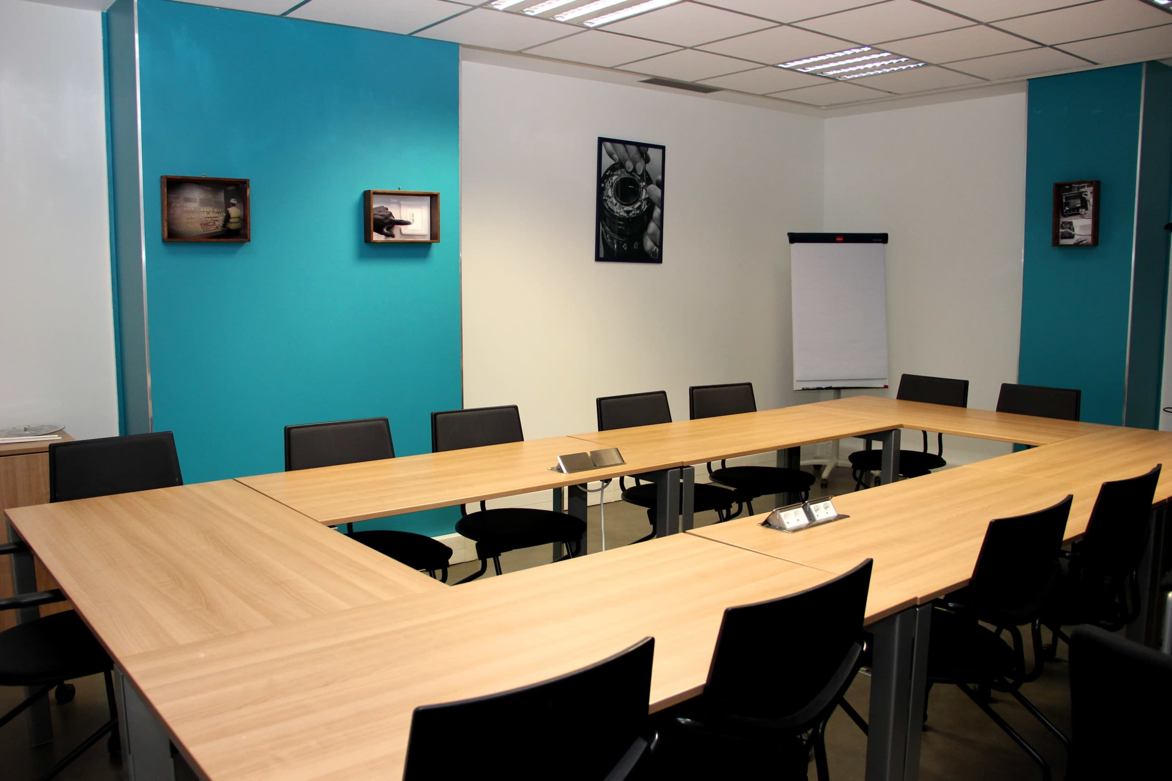 A conference room features a large, rectangular wooden table surrounded by black chairs, with blue accent walls and framed pictures hung on the walls.
