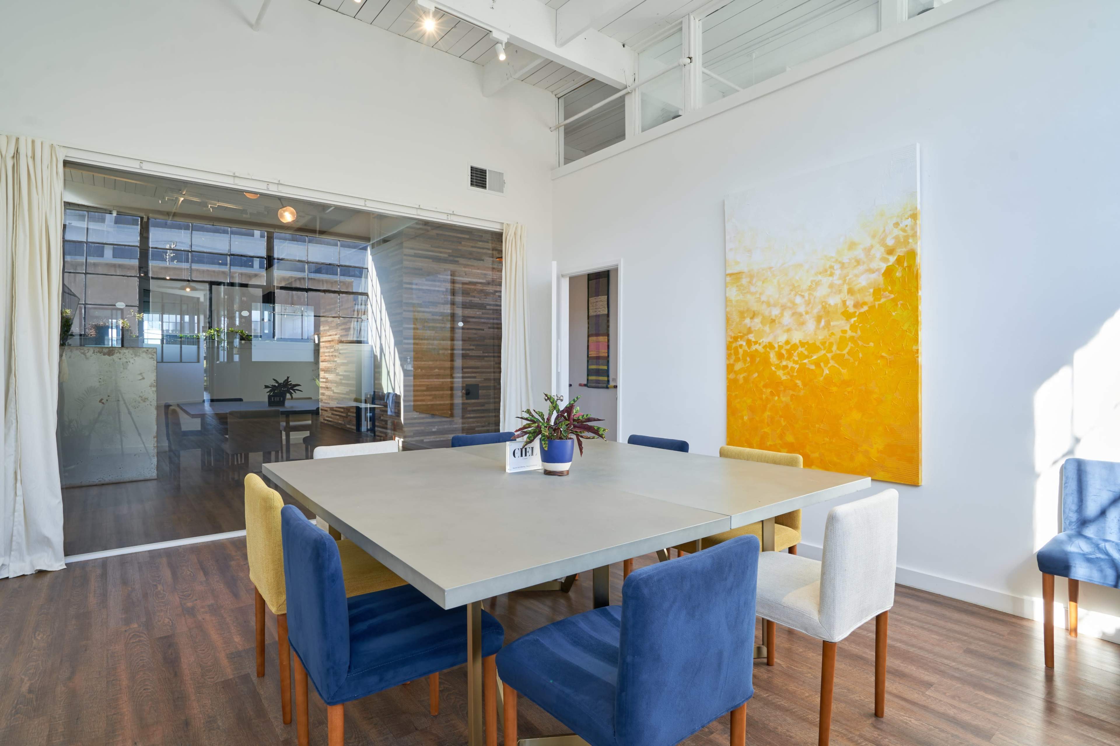A bright meeting room features a large concrete table surrounded by colorful chairs and a large abstract yellow painting on the wall.