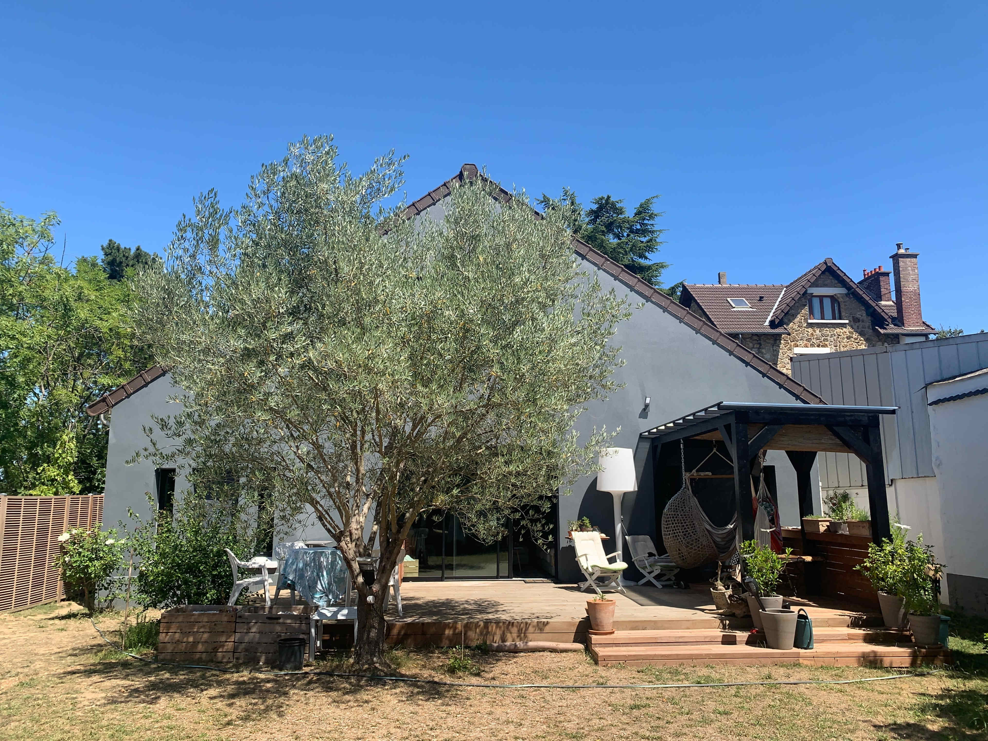 A modern house with a spacious wooden deck surrounded by plants and trees, set against a clear blue sky.