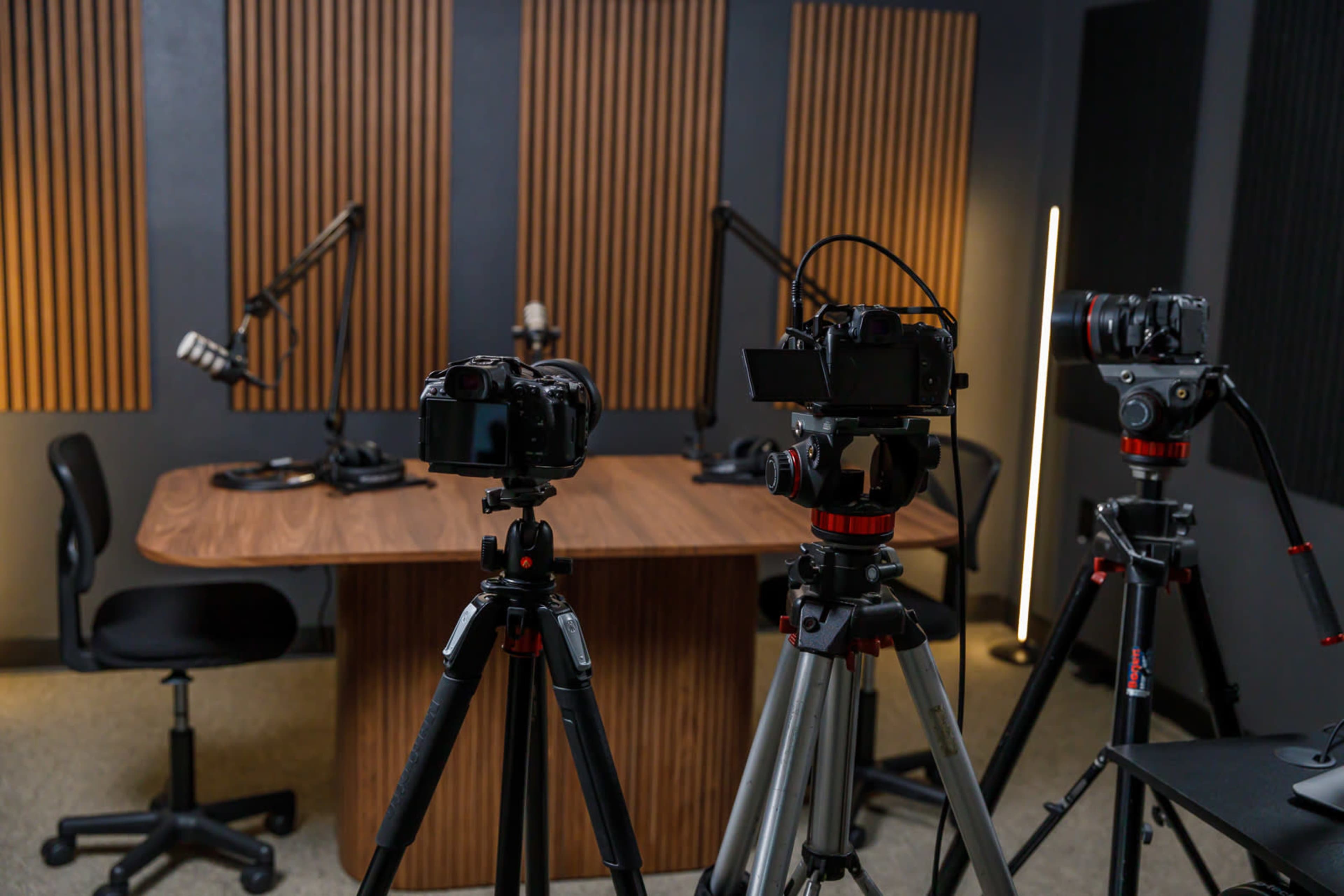 A studio setup features multiple cameras on tripods facing a wooden table with microphones and chairs in an acoustically treated room.