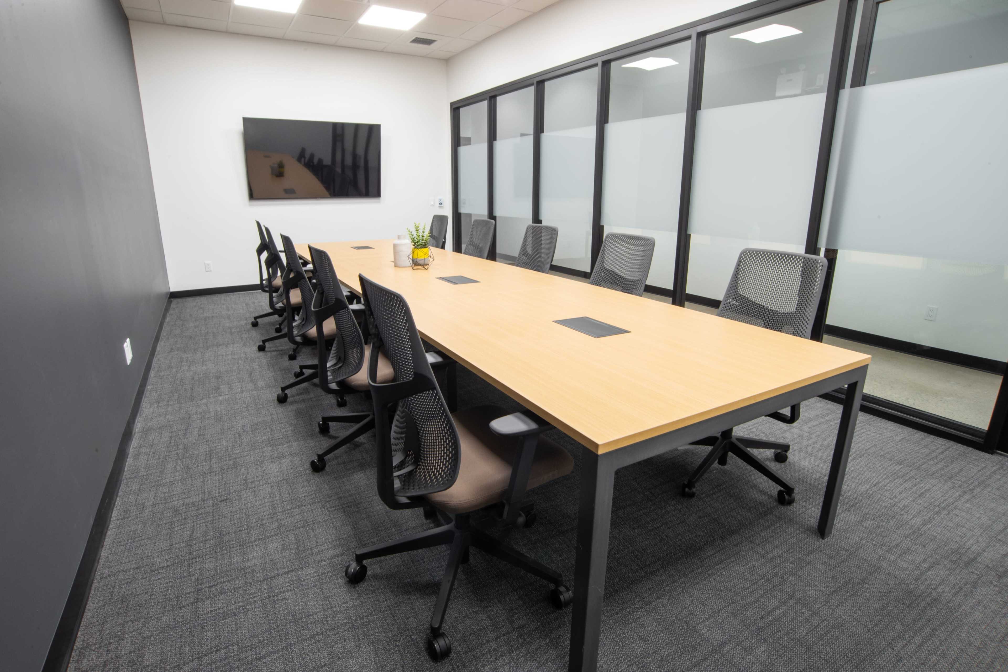 The image shows a modern conference room with a long wooden table surrounded by ergonomic chairs and a wall-mounted screen.