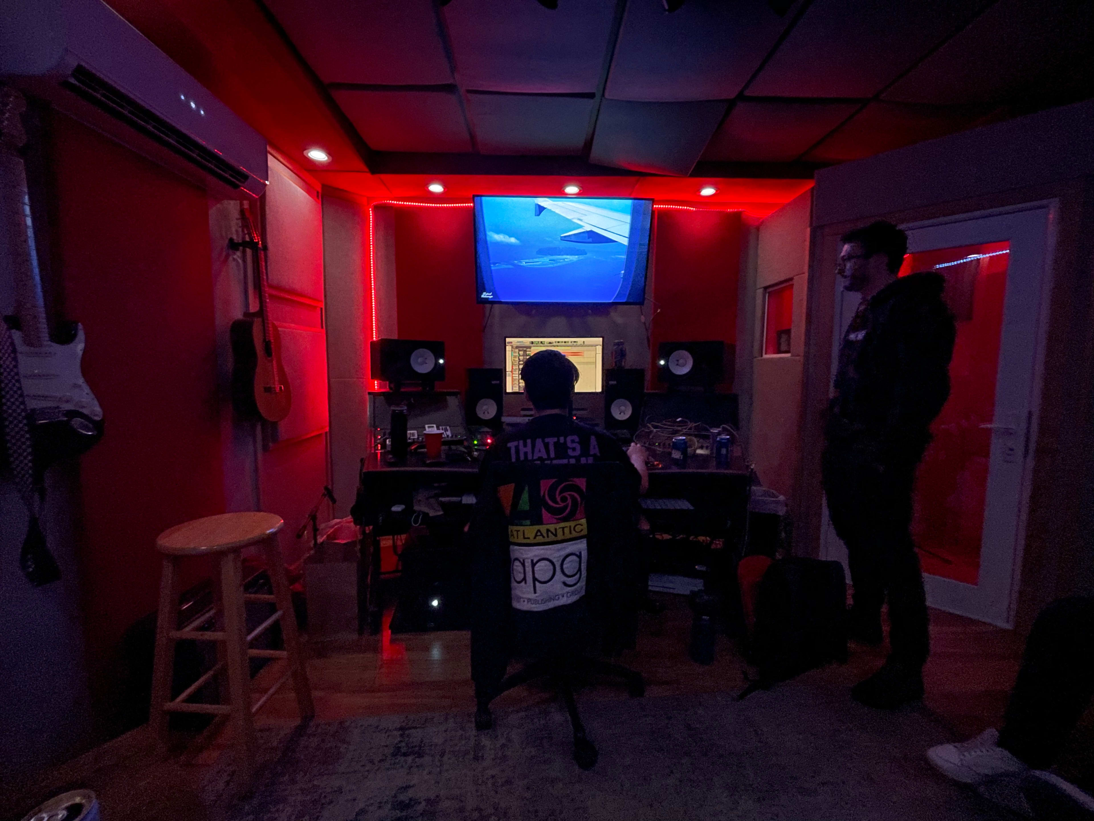 A person sits at a studio desk with audio equipment, while another person stands nearby in a dimly lit room featuring red lighting and sound monitors.