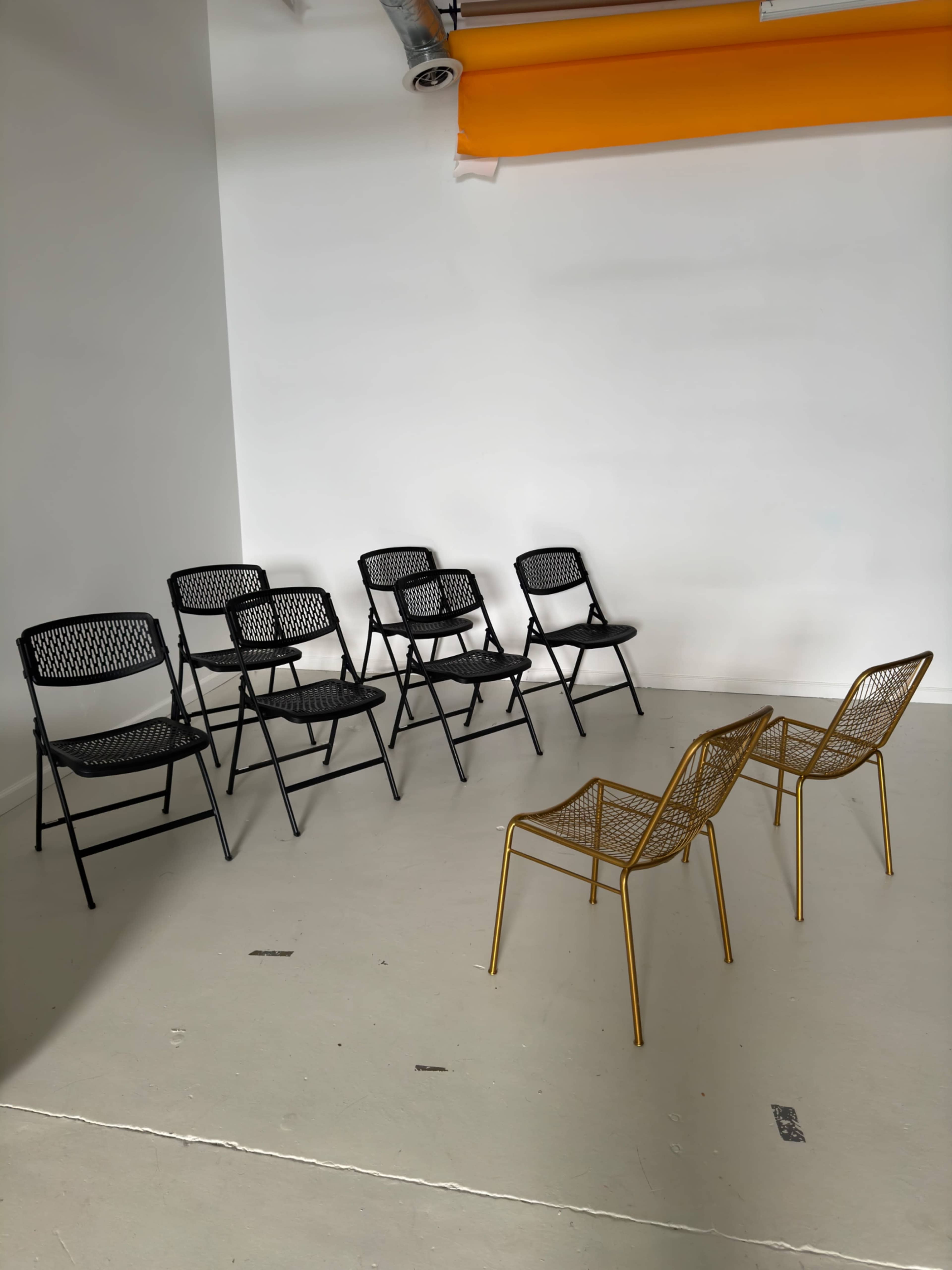 A group of six black folding chairs is arranged against a wall, facing two gold wire chairs in a minimalist room with a white backdrop.