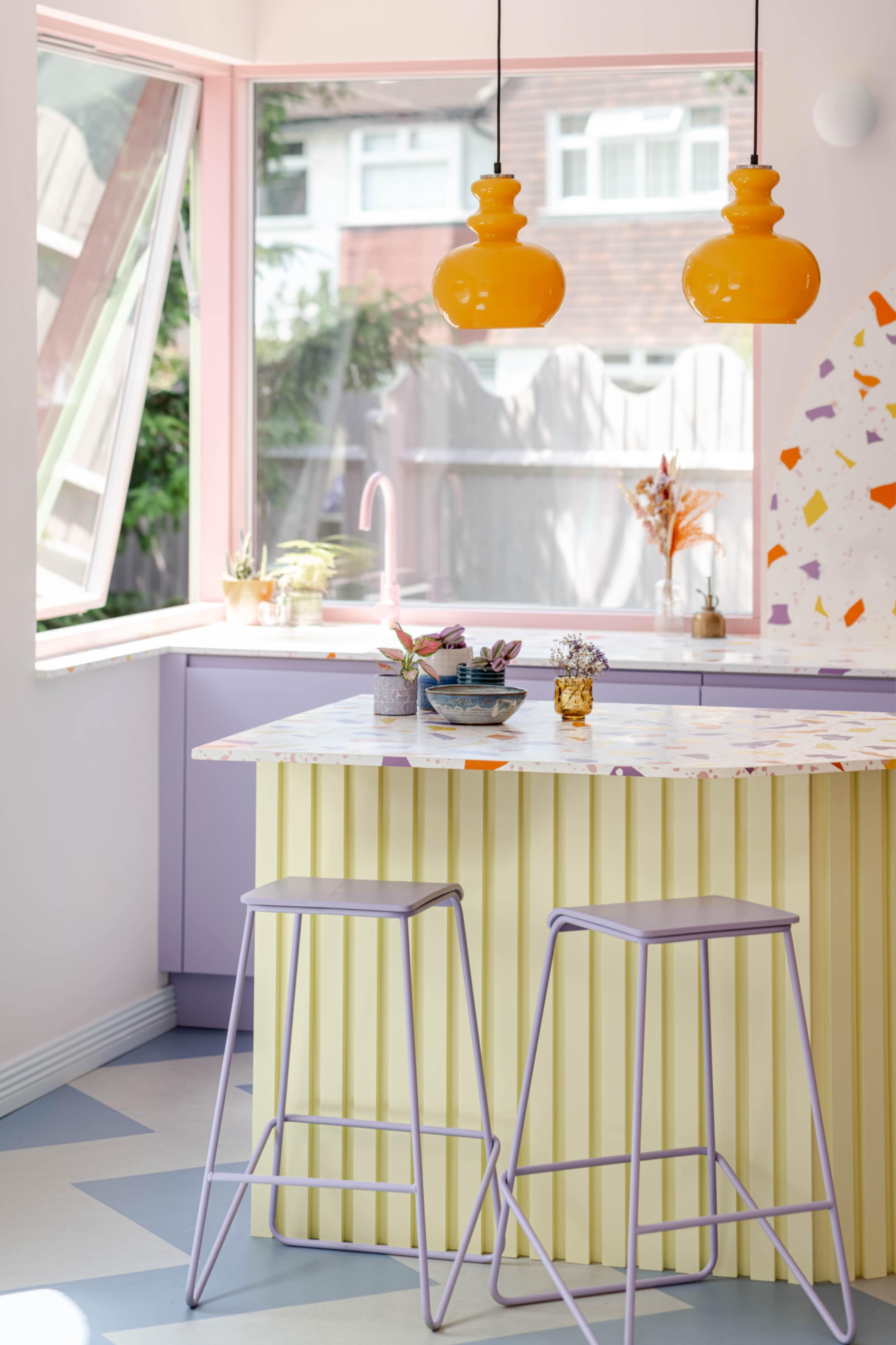 A modern kitchen features a yellow ribbed island with purple bar stools, large windows, and colorful pendant lights above.