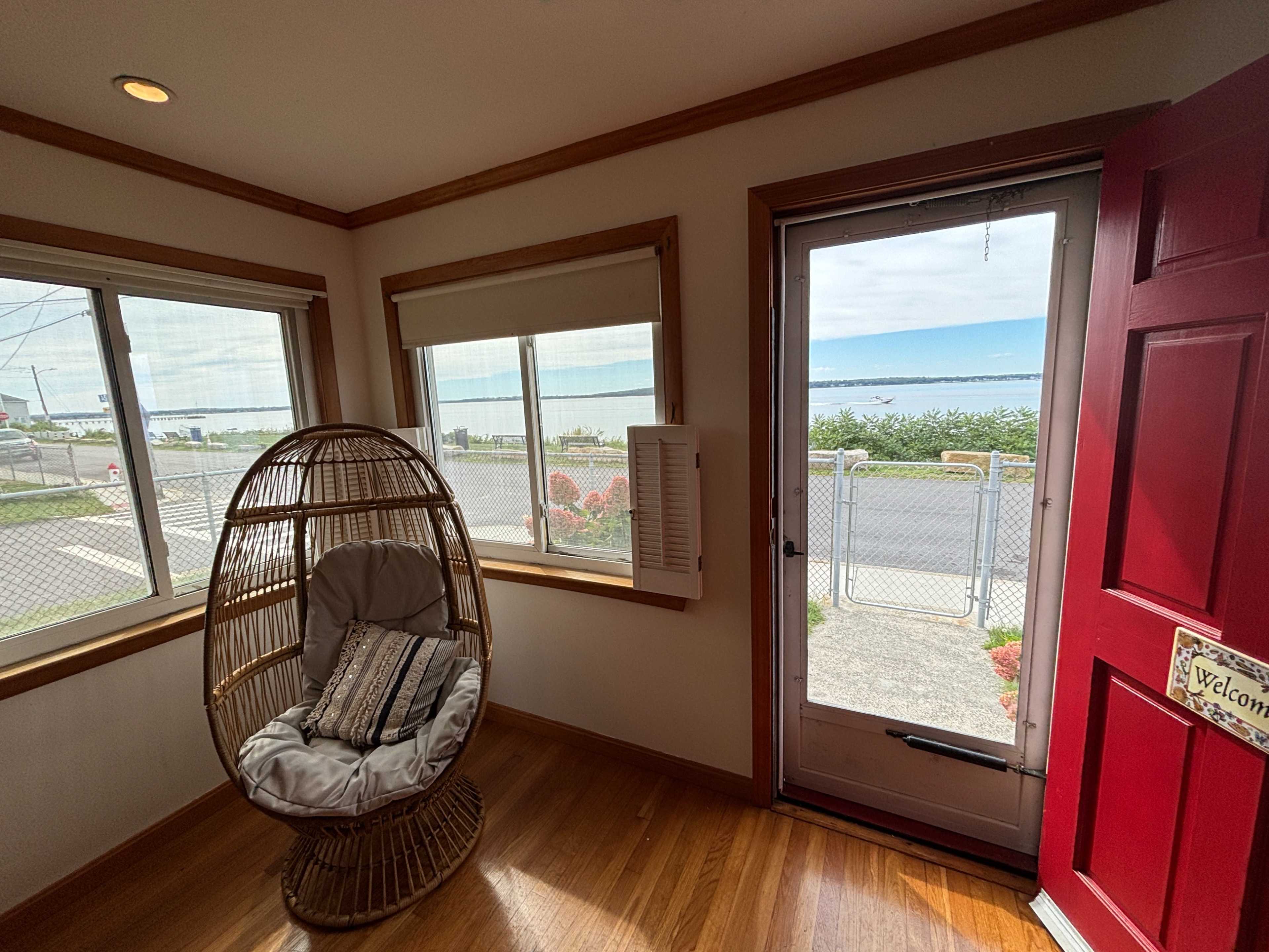 A wicker chair is positioned by a door with a red frame, overlooking a view of the water and nearby landscaping.
