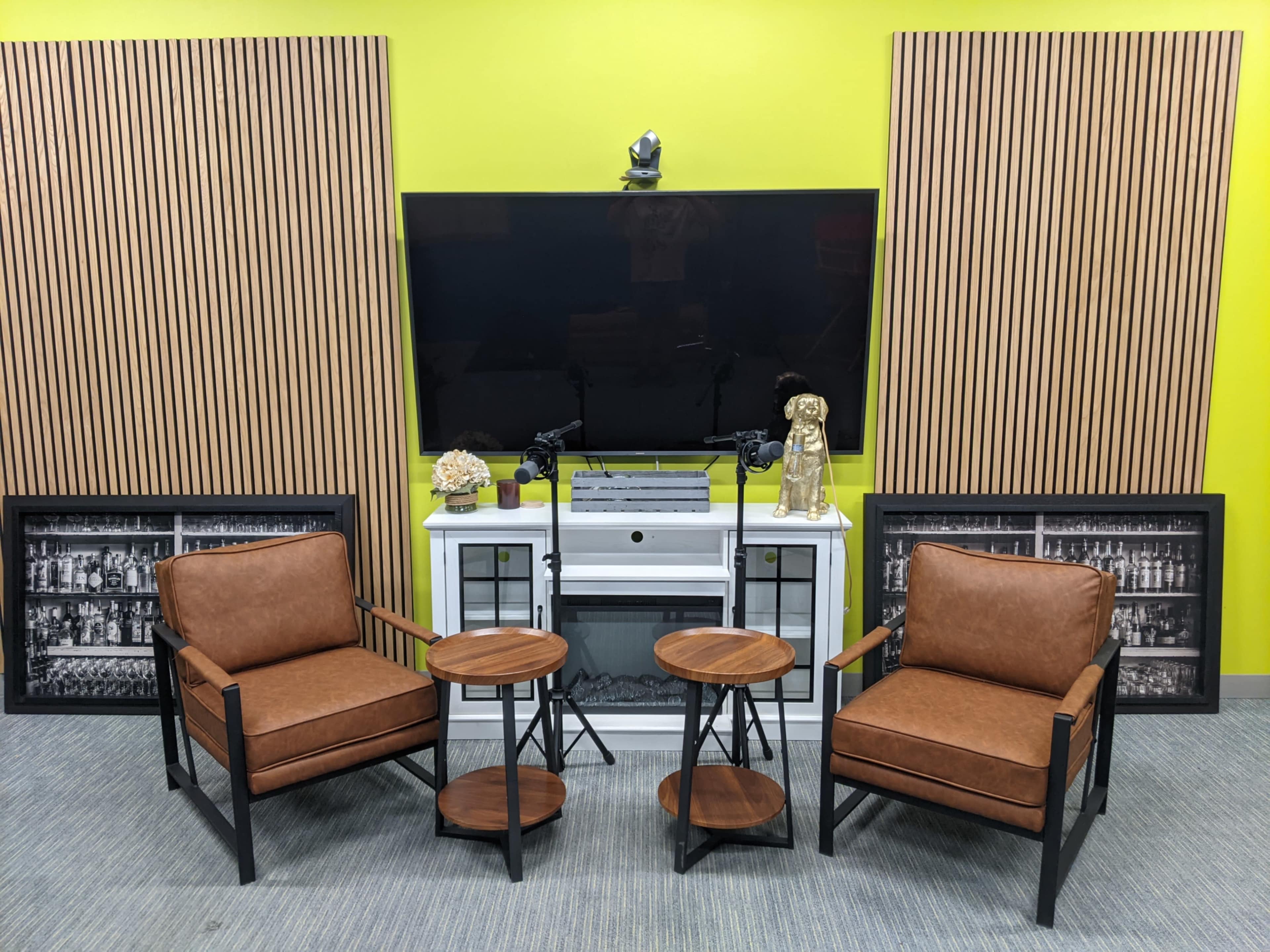 The image shows a modern seating area with two brown chairs, two round wooden tables, a white cabinet, and a large TV mounted on a lime green wall, flanked by wooden paneling.