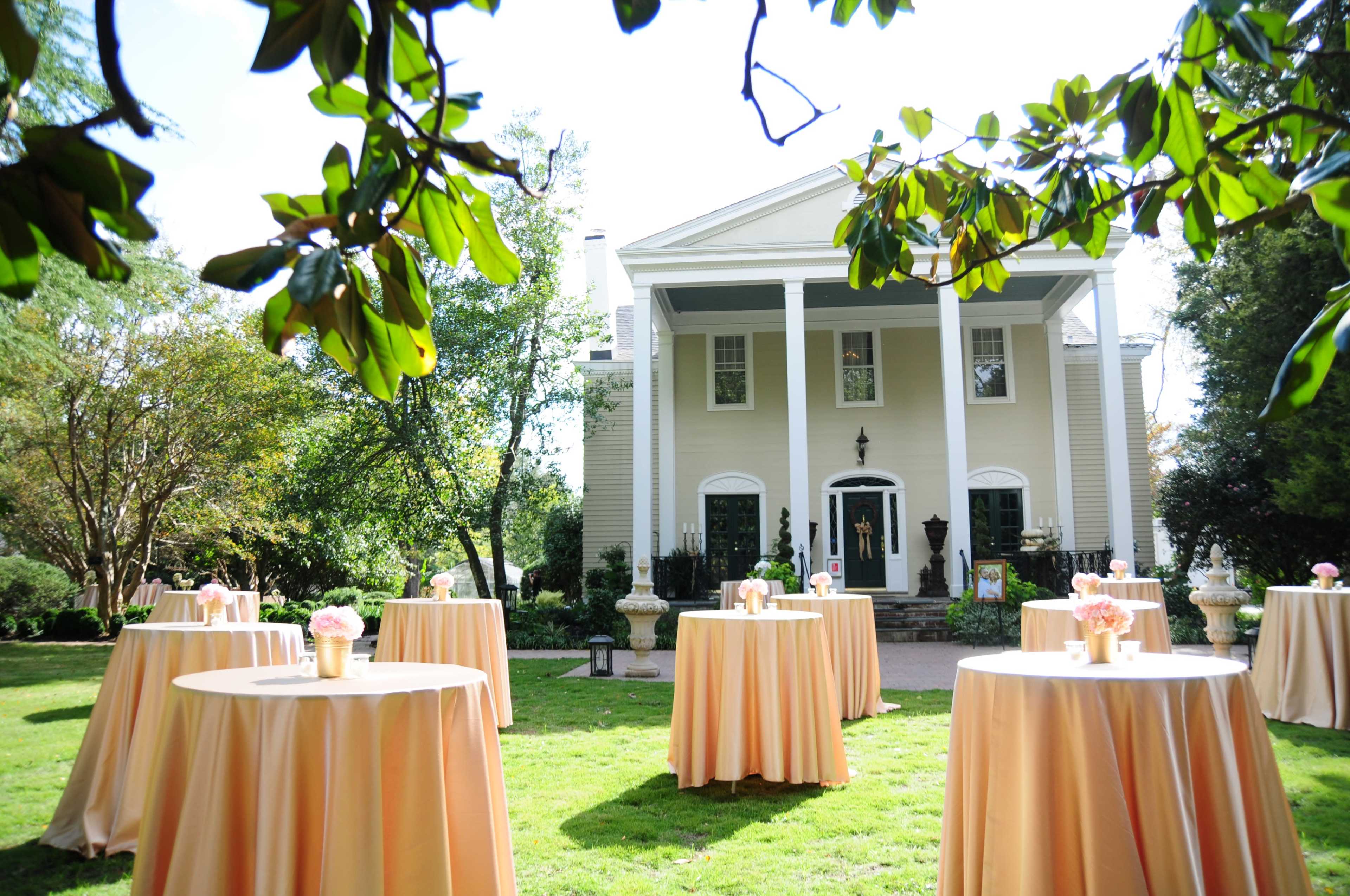 An elegant outdoor setting features several round tables covered with light orange tablecloths in front of a spacious house.