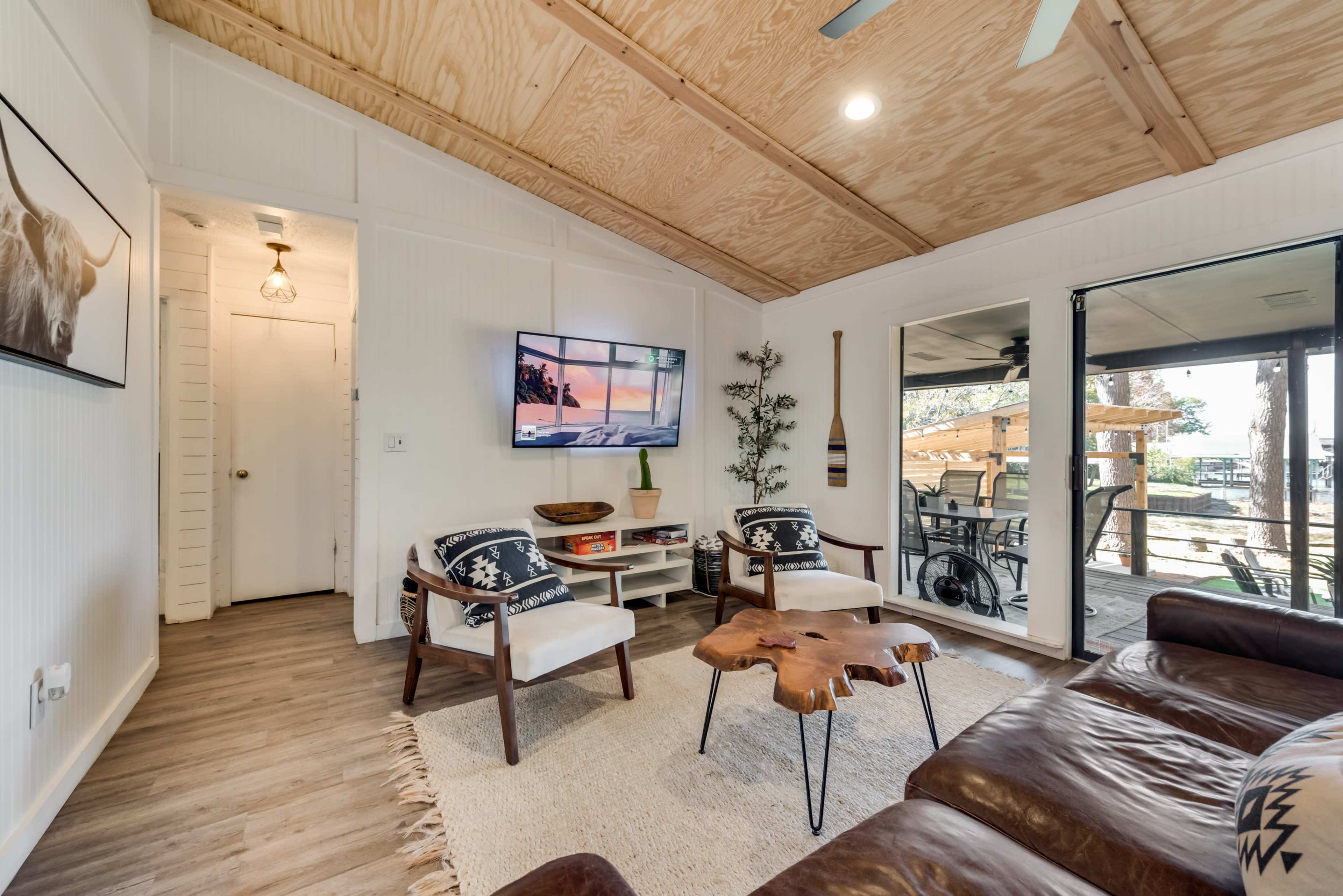 A cozy living room with a wooden ceiling, two armchairs, a coffee table, and a TV mounted on the wall, leading to a deck area outdoors.