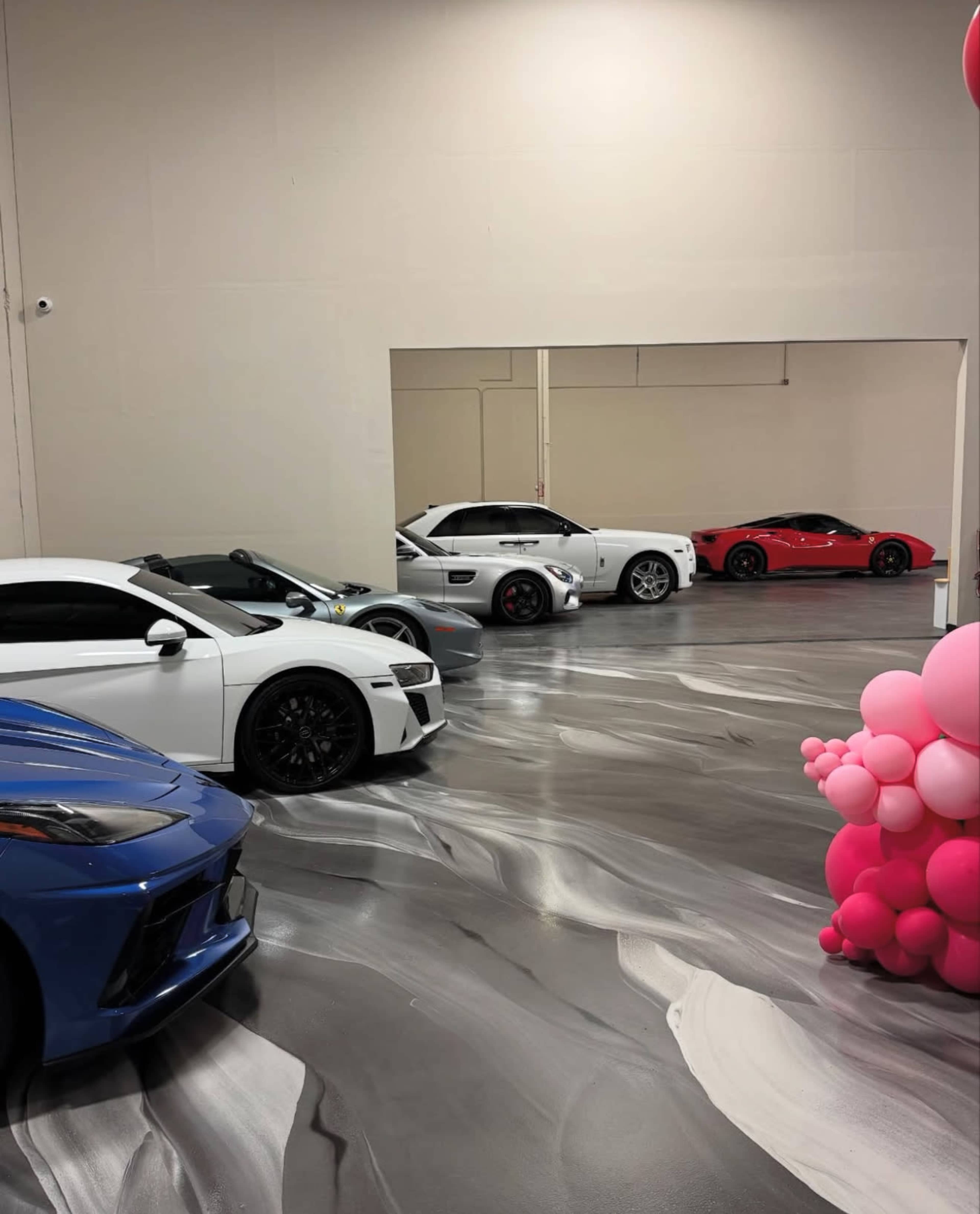 The image shows a garage with five distinct cars parked on a marbled gray floor, with a display of pink balloons on the right side.