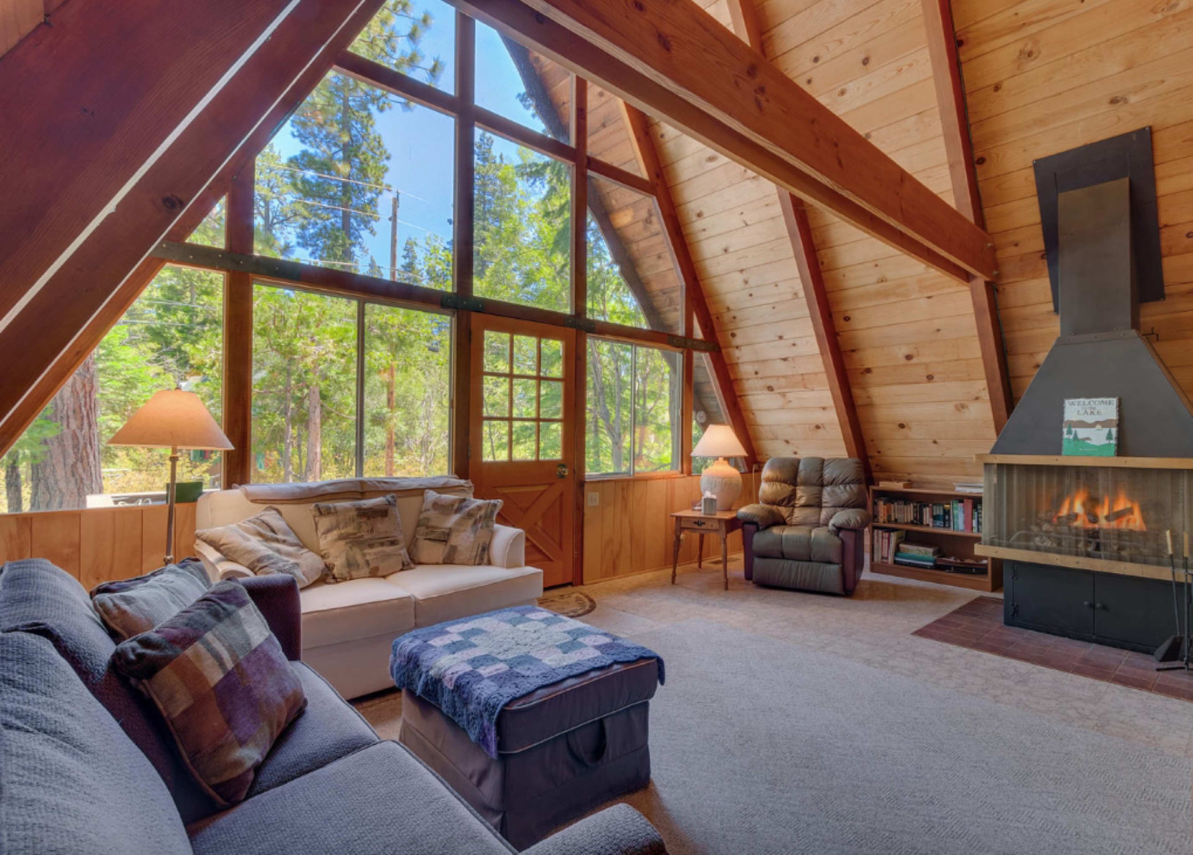 A cozy cabin interior features large windows, wooden beams, a fireplace, and comfortable seating.