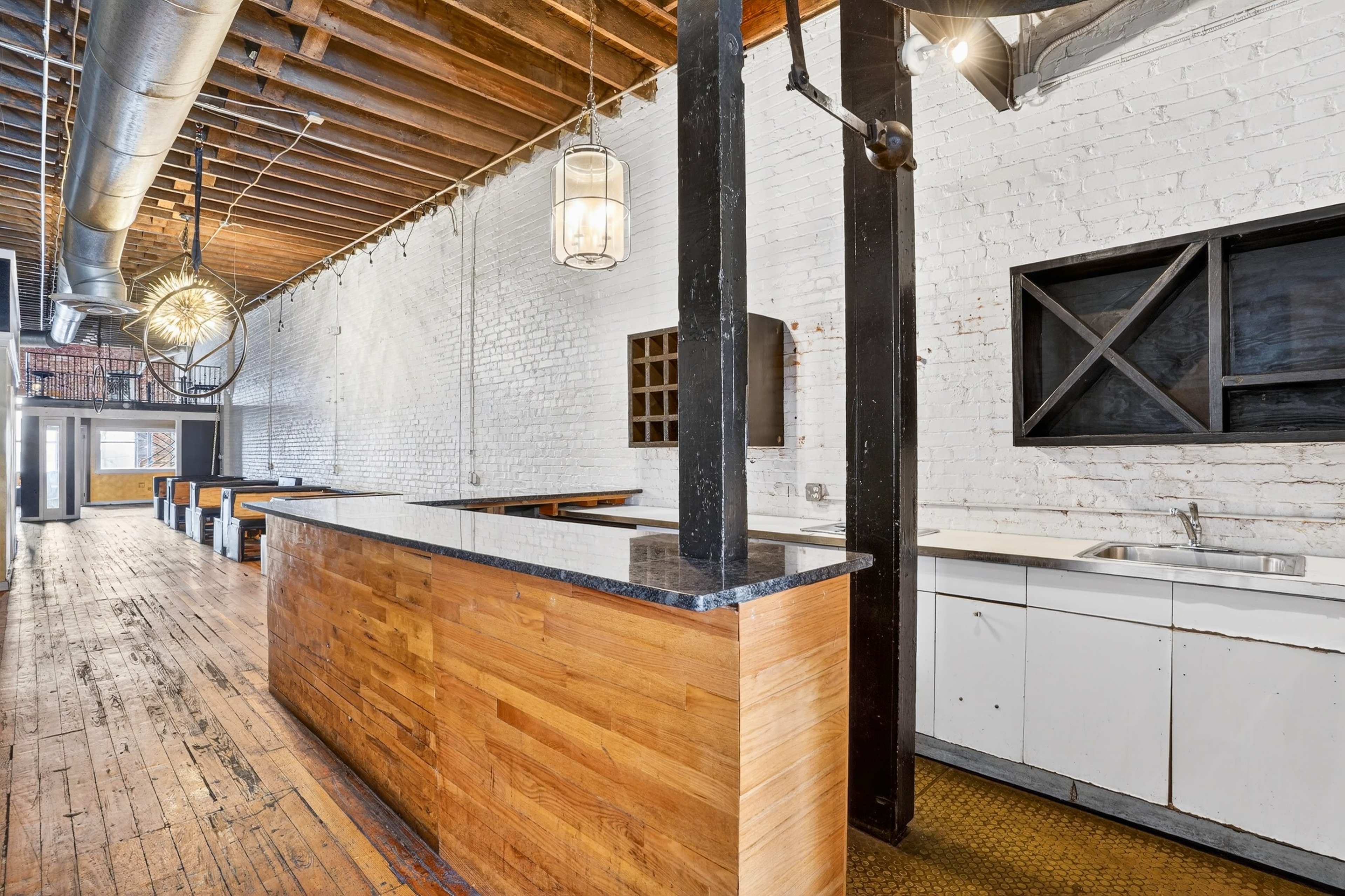 The image shows a spacious, modern kitchen area with a wooden bar, stainless steel fixtures, and exposed brick walls under a high ceiling with wooden beams.