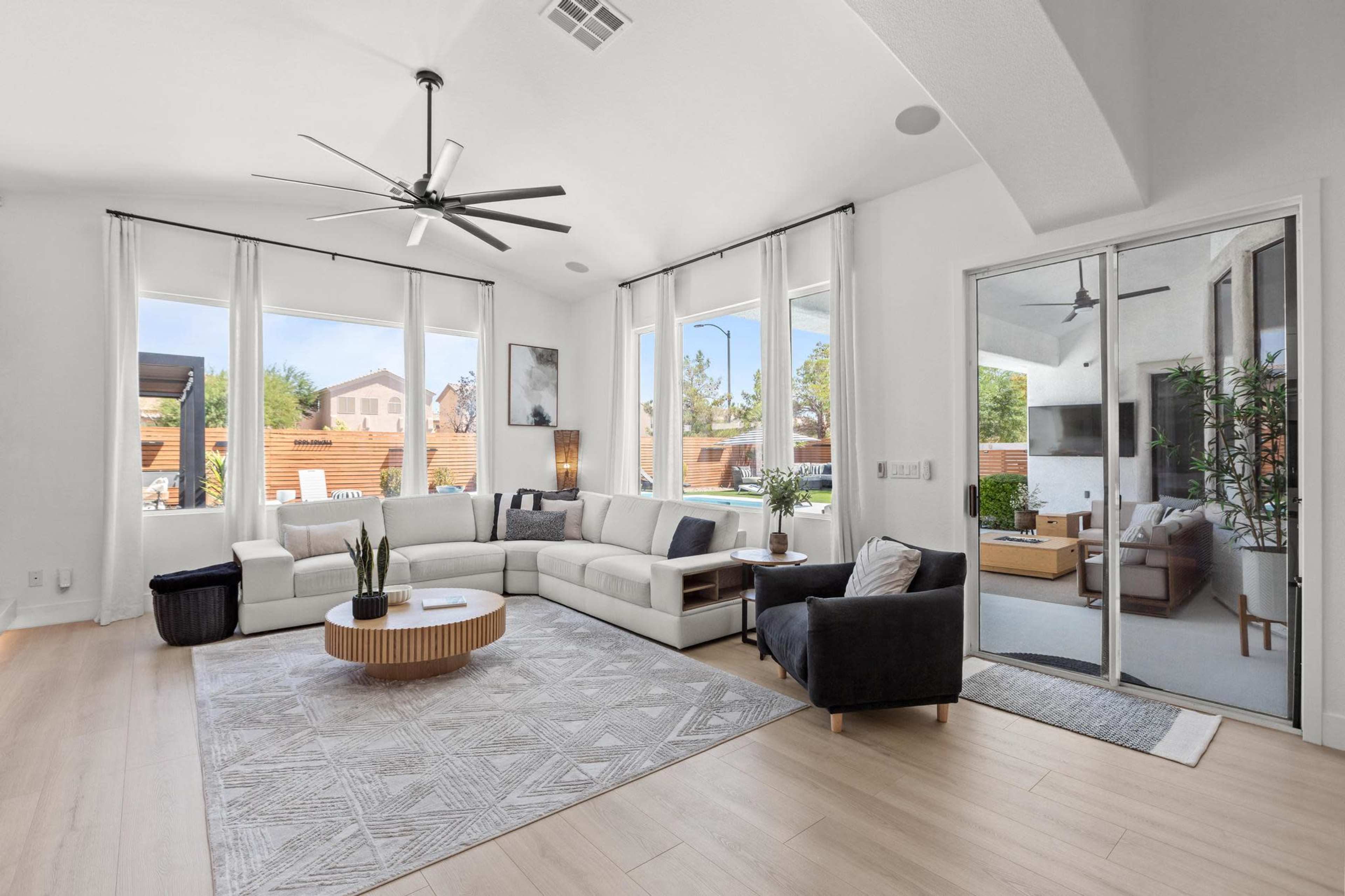 The image shows a spacious living room with large windows, a sectional sofa, a coffee table, and access to an outdoor patio area.