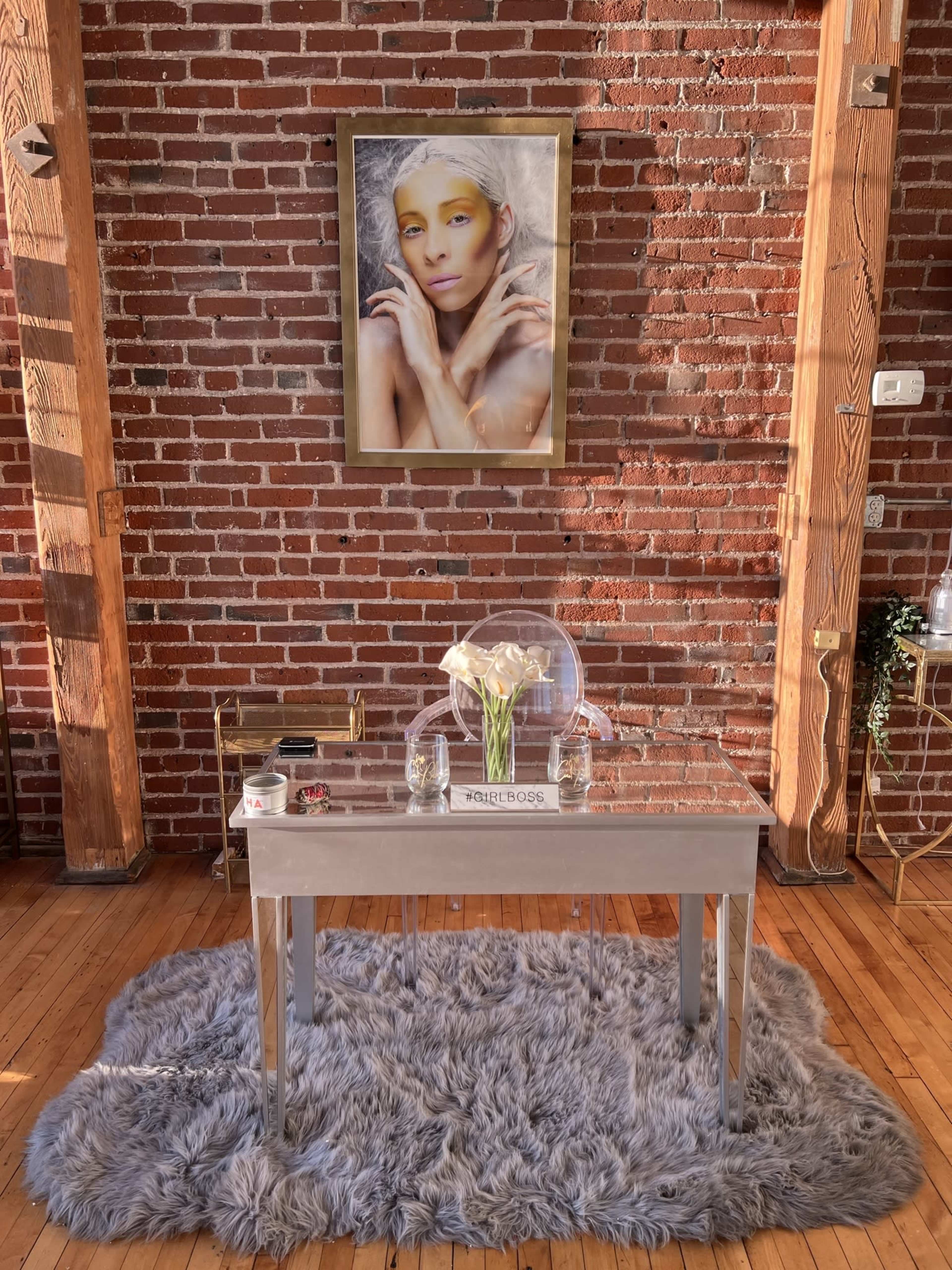 A small silver table is placed on a gray rug in front of a brick wall, adorned with a framed portrait of a person hanging above it.