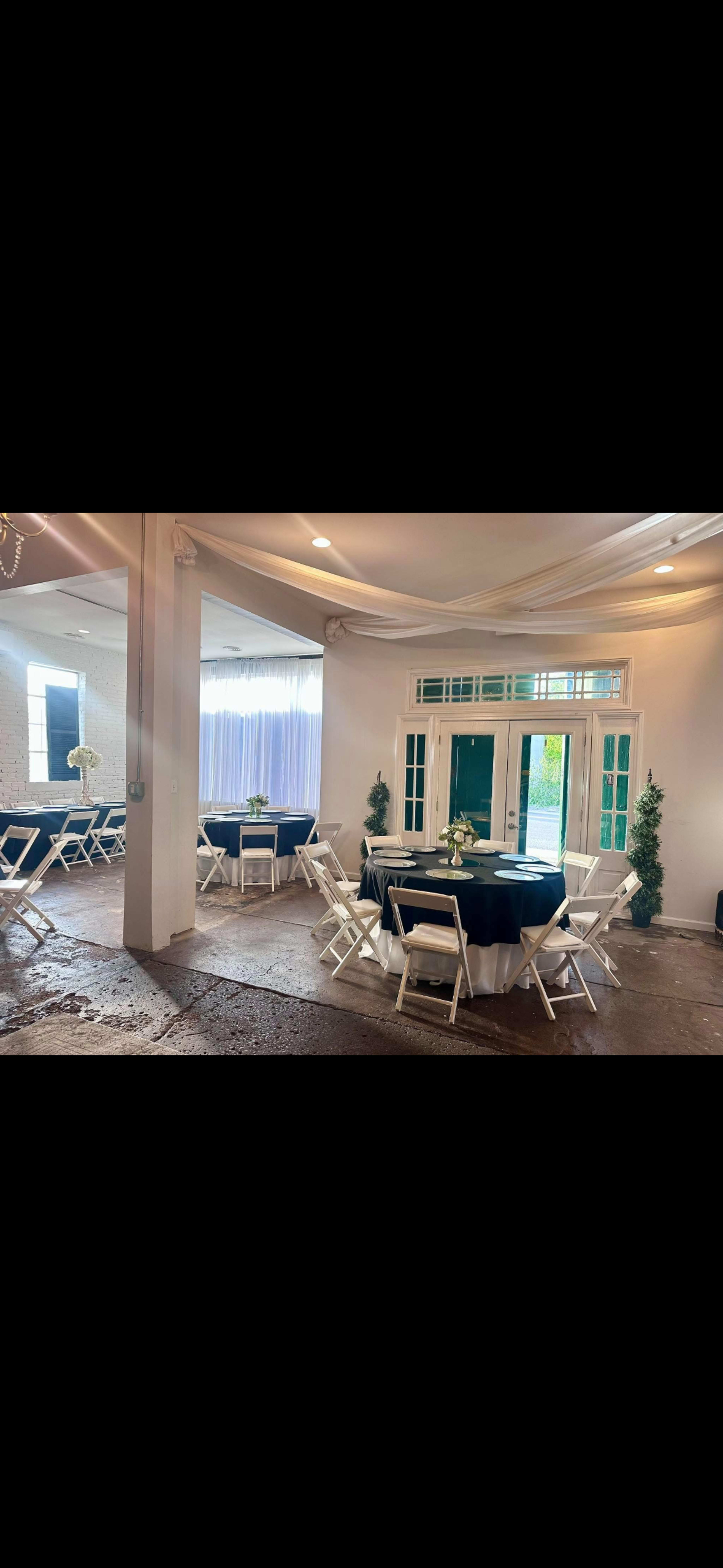 The image shows a decorated indoor venue with round tables covered in navy blue cloth, white folding chairs, and potted plants, surrounded by large windows and sheer curtains.