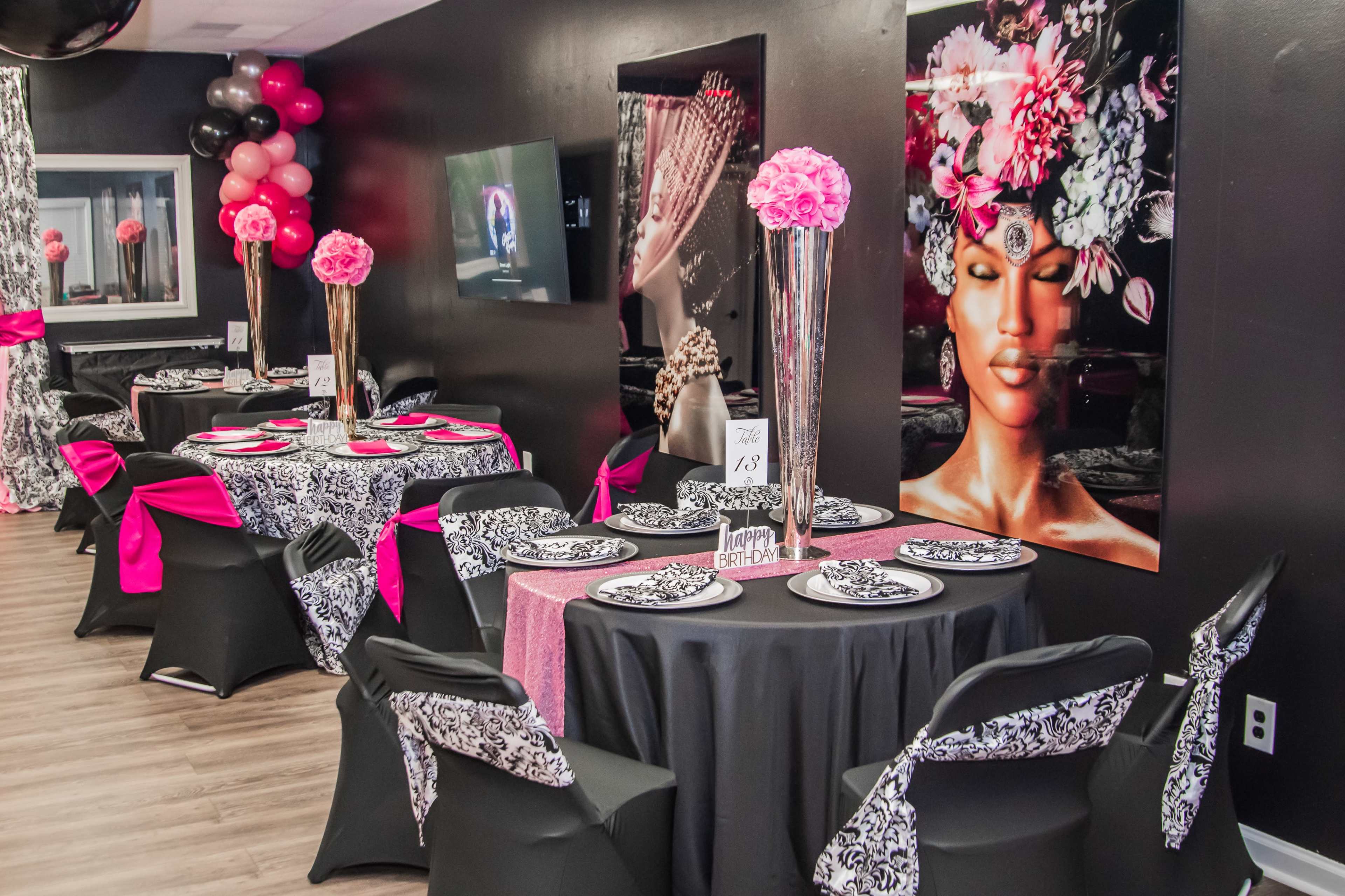 A stylish event space featuring tables with black and pink table settings, adorned with floral centerpieces and decorative balloons.