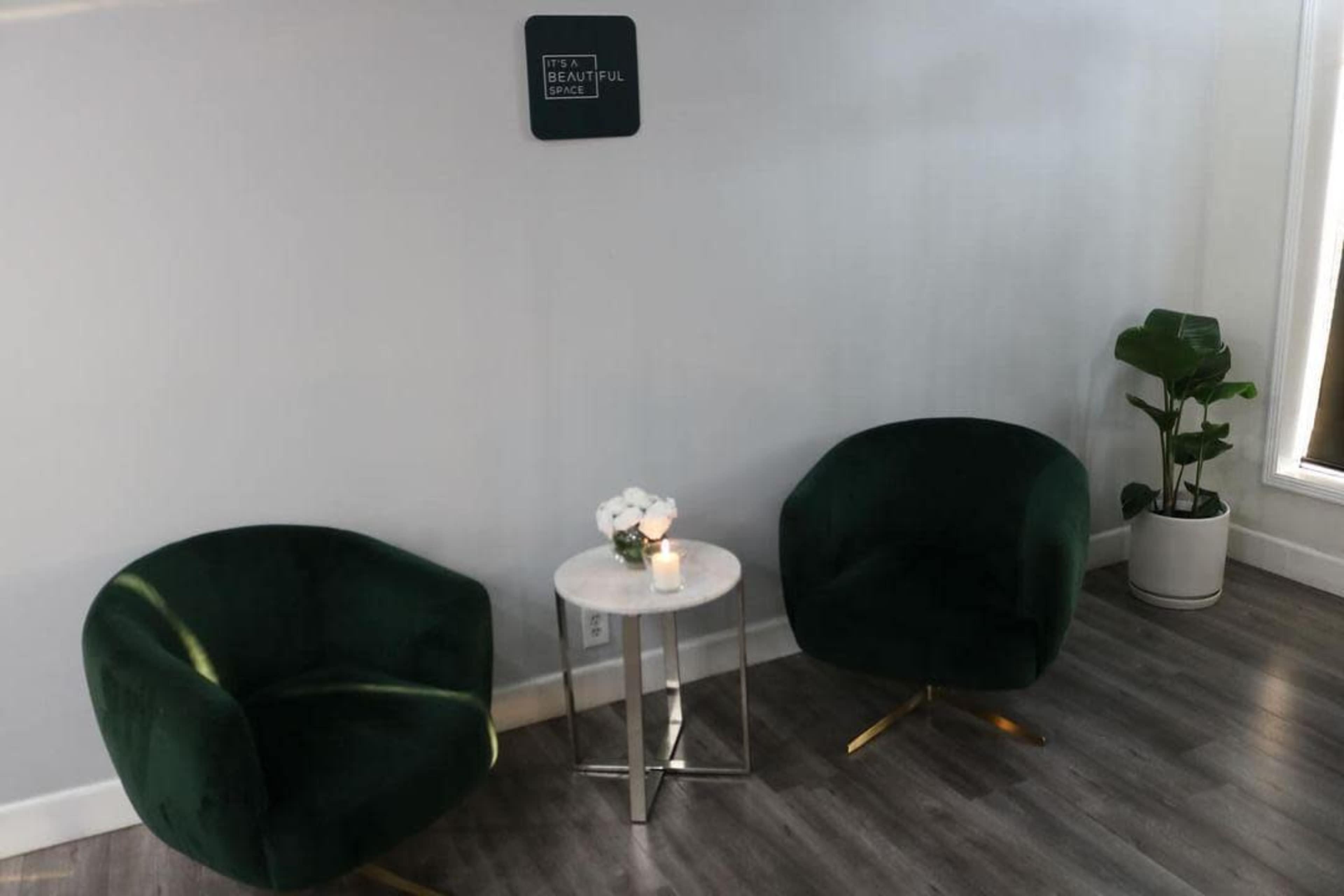 The image shows a cozy corner of a room featuring two green velvet chairs, a small round table with a candle, and a potted plant against a light grey wall.