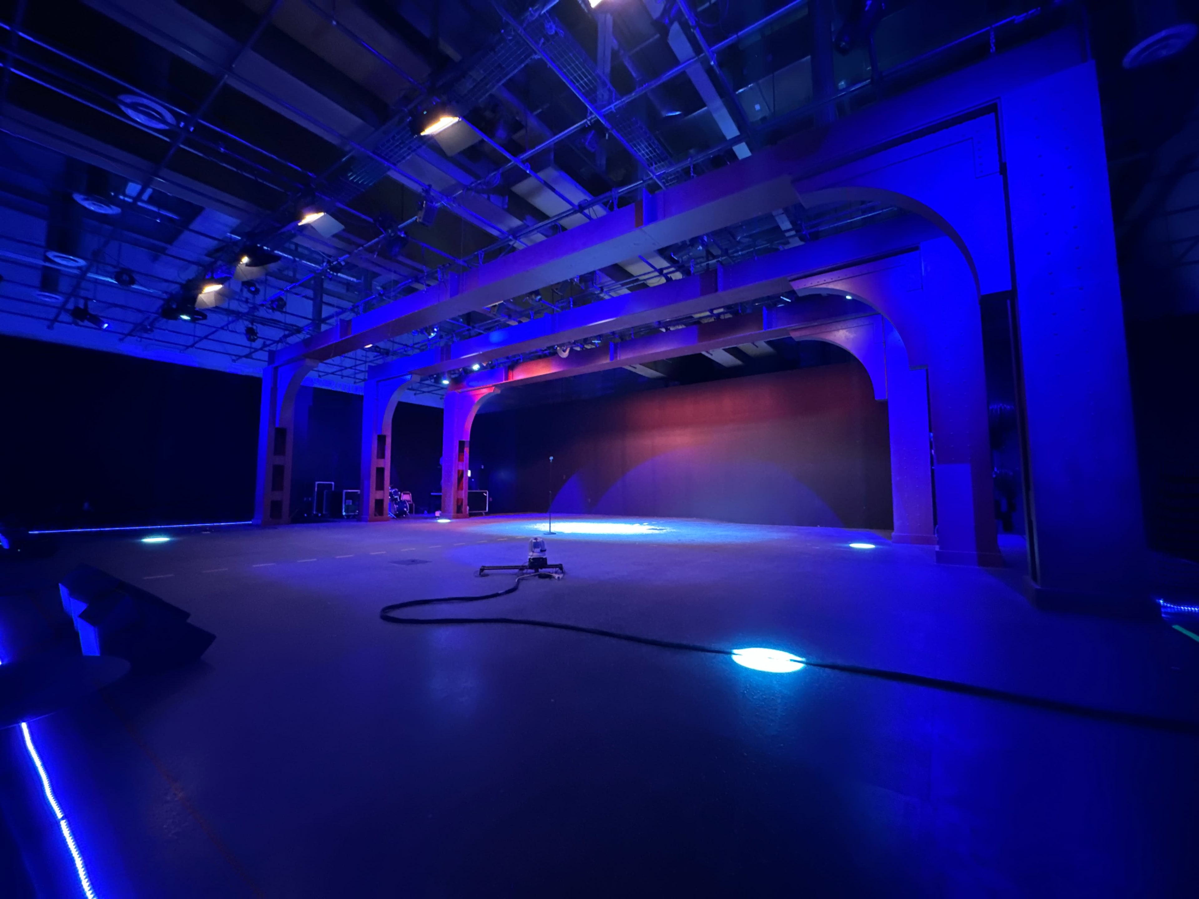 A large, industrial space is illuminated by blue and purple lights, featuring arched supports and a clear stage area.