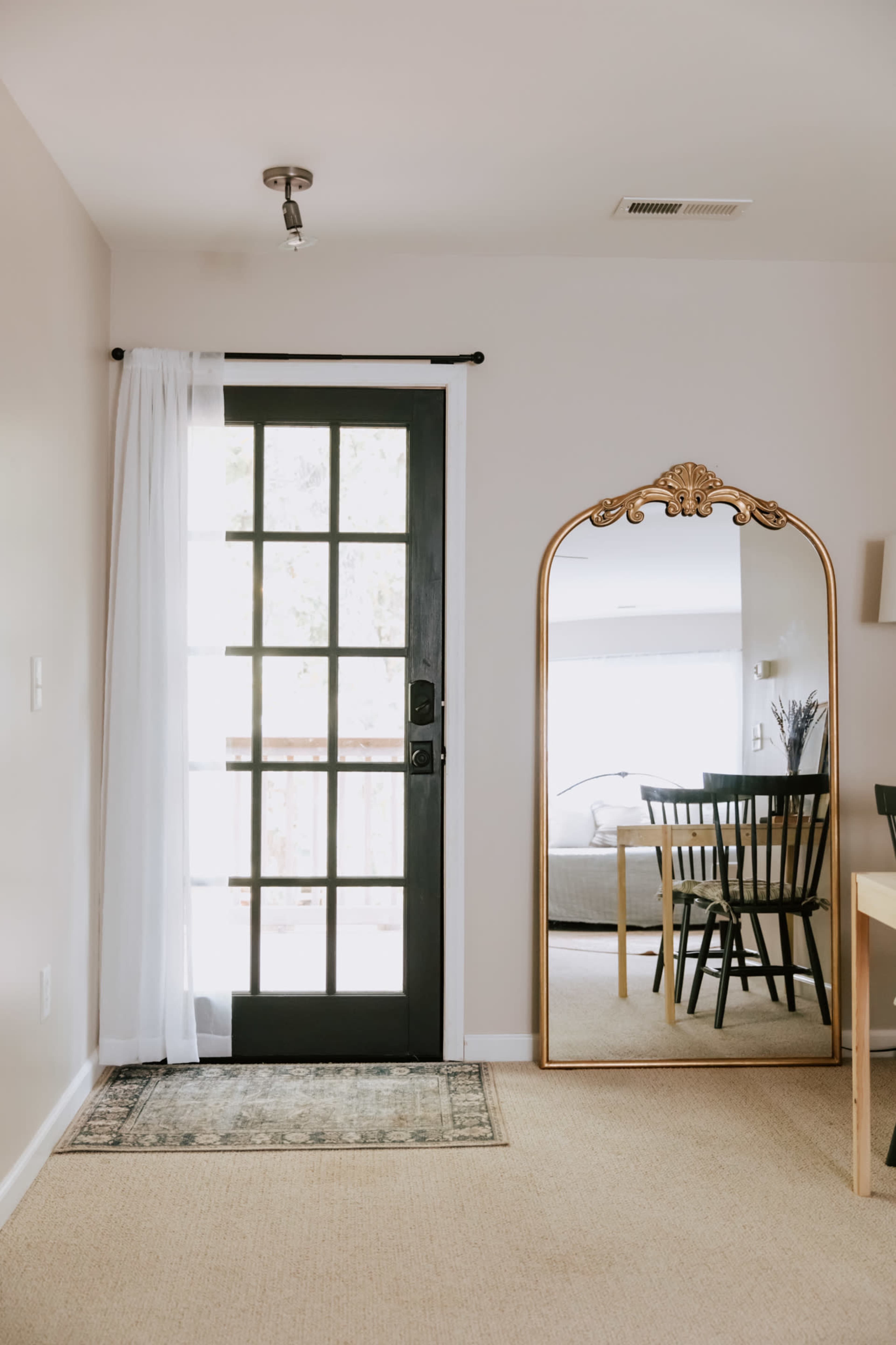 A black door with glass panes opens into a room featuring a large, ornate mirror, a dining table, and chairs.