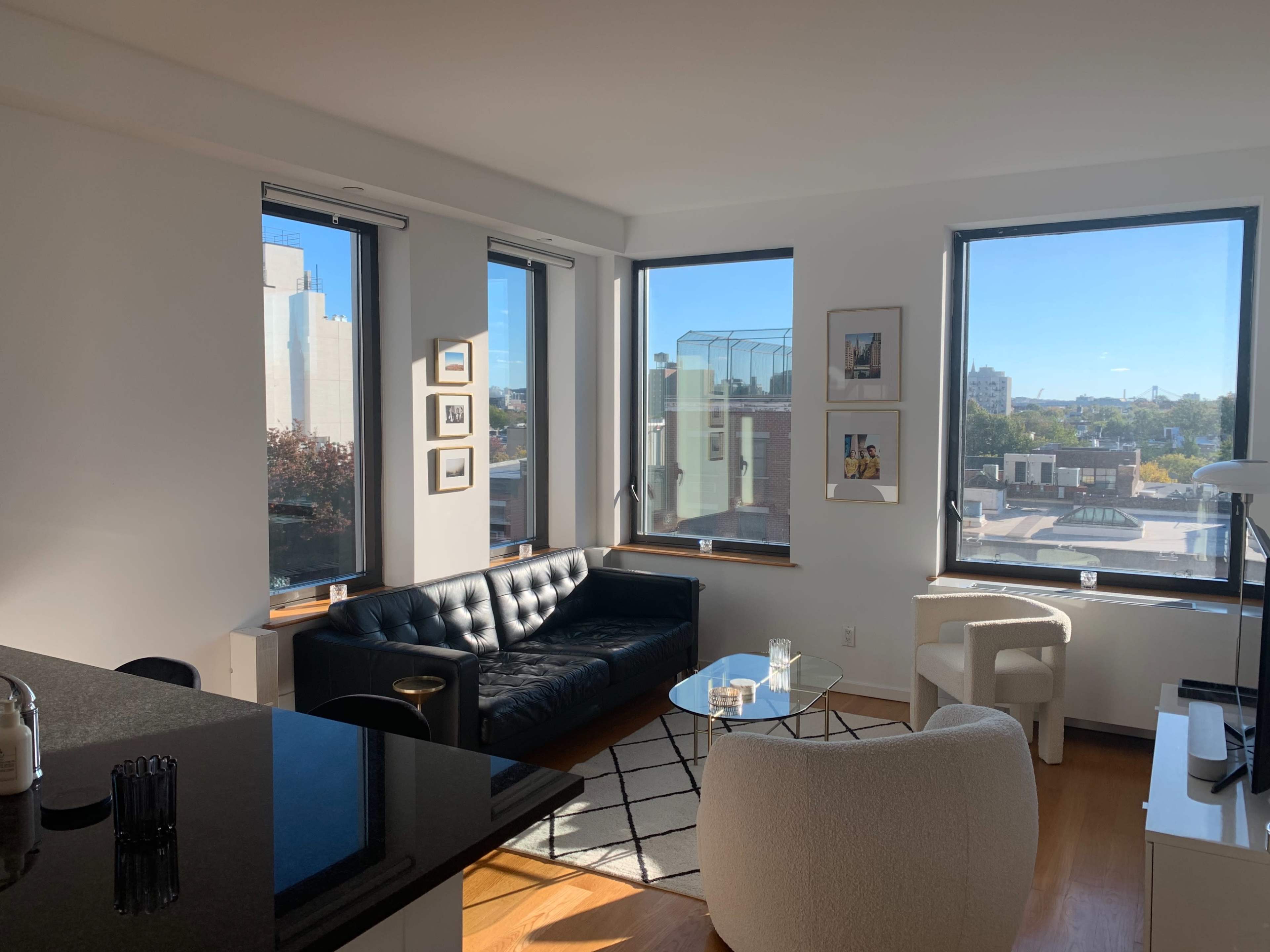 A modern living room with a black sofa, glass coffee table, and large windows that provide natural light and a view of the city.