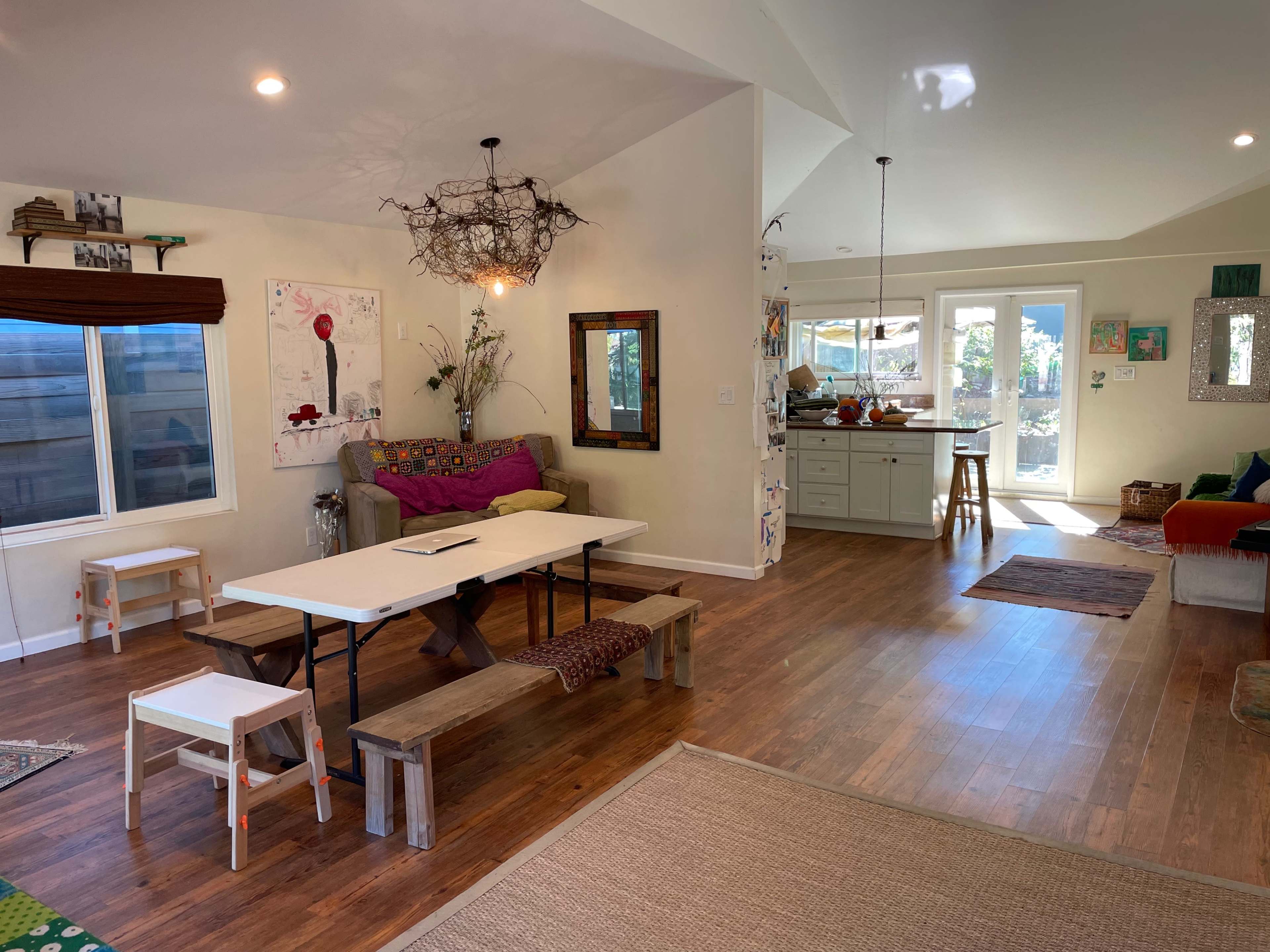 The image shows a bright, open living space with a dining table, a seating area, and a kitchen visible in the background.