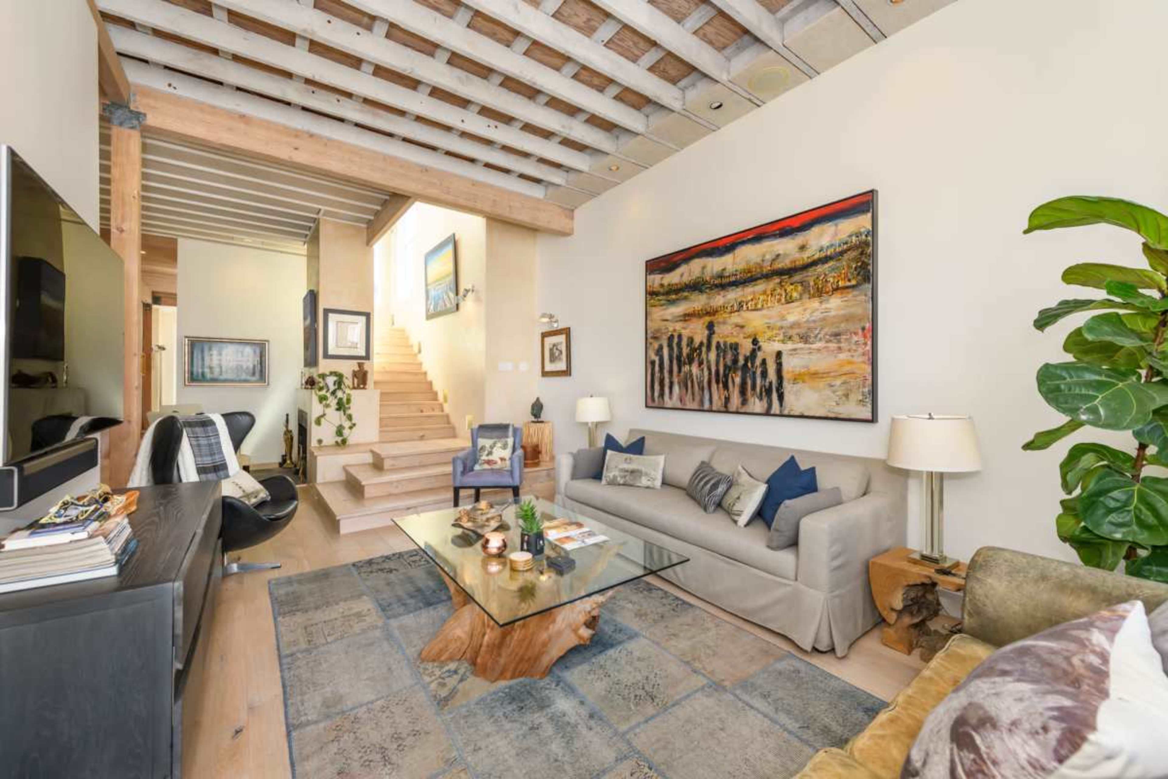 The image features a modern living room with a cozy sofa, a glass coffee table, and a staircase leading to an upper level, adorned with artwork and numerous plants.