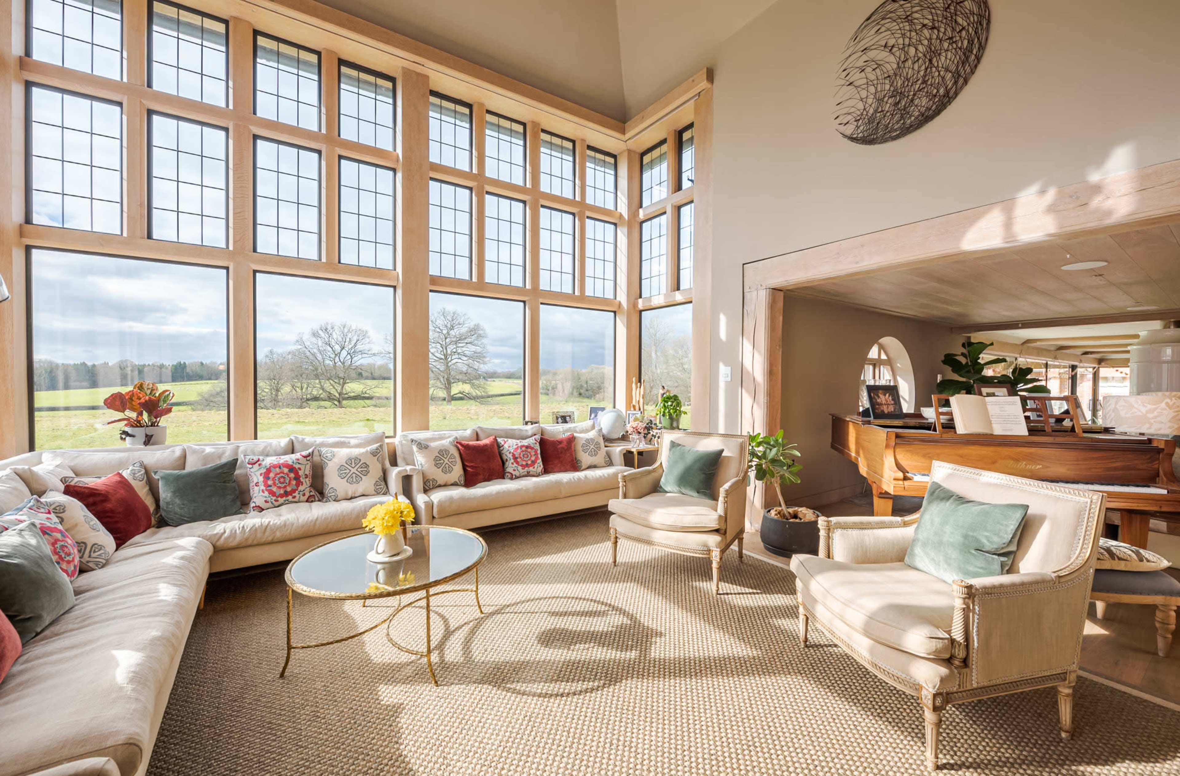 The room features large windows overlooking a grassy landscape, with a light-colored sofa adorned with decorative pillows and two armchairs arranged around a circular coffee table.