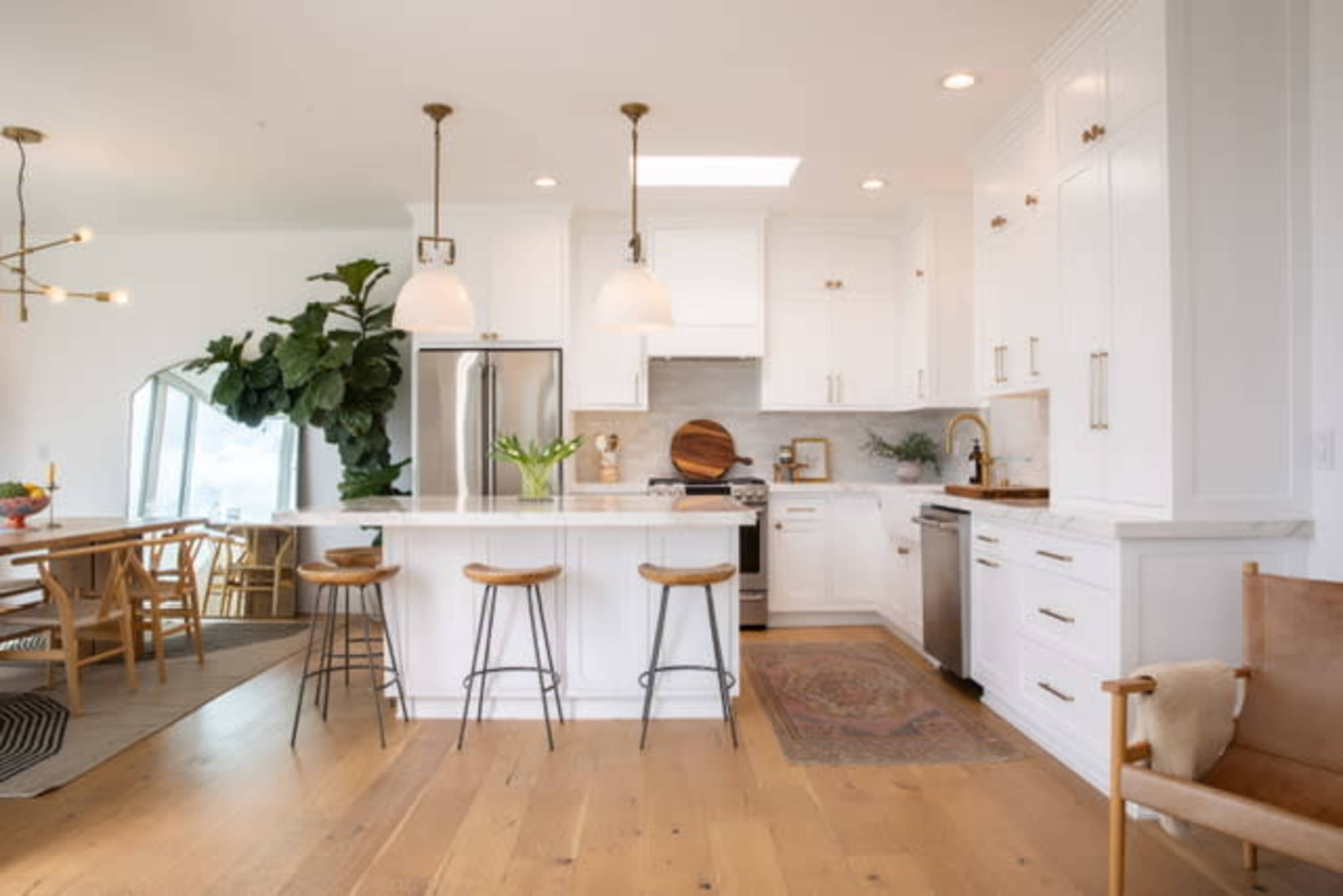 A modern kitchen with white cabinetry, a central island with three wooden stools, and large windows providing natural light.