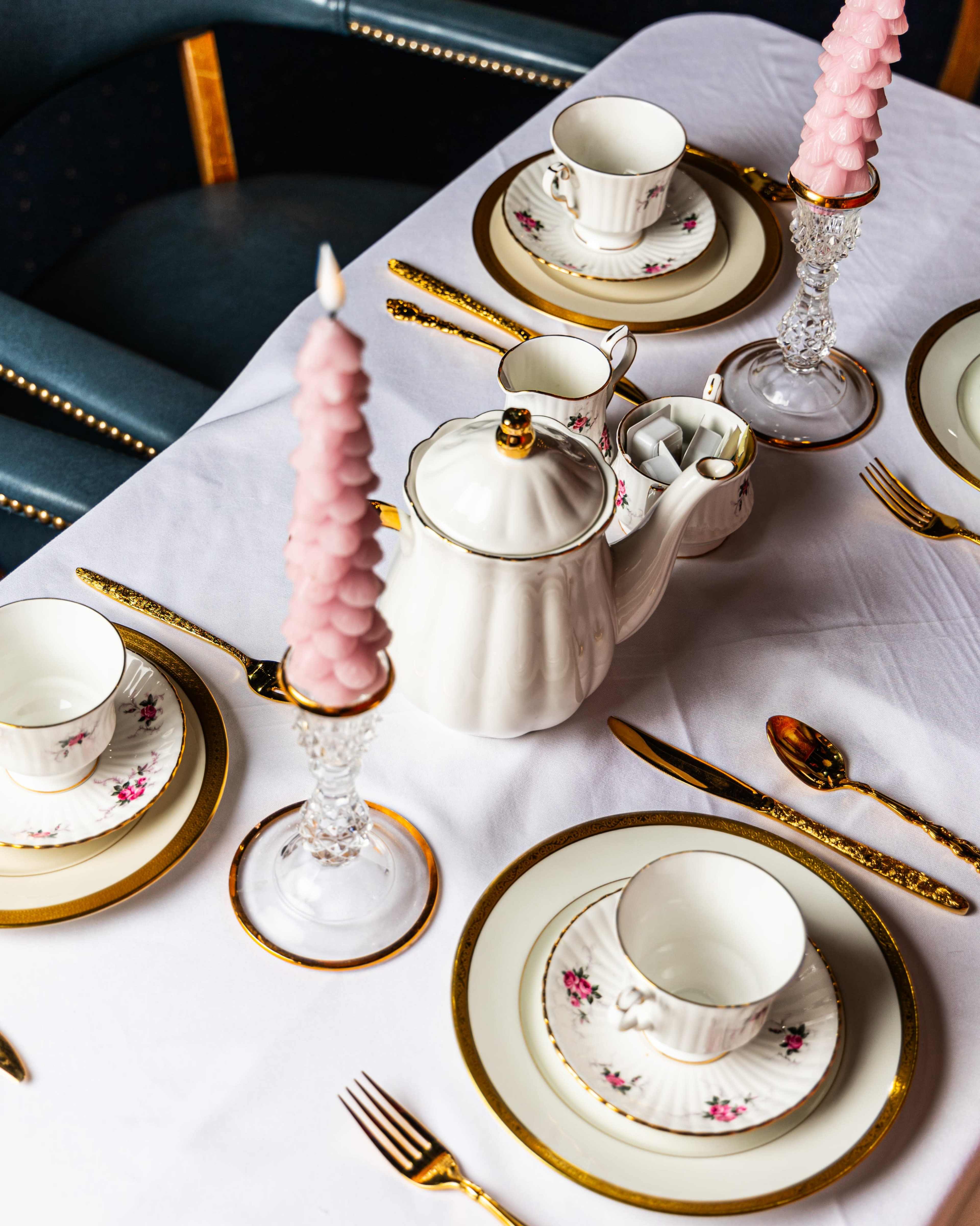 A table is elegantly set with fine china, a teapot, crystal candle holders, and pink taper candles on a white tablecloth.