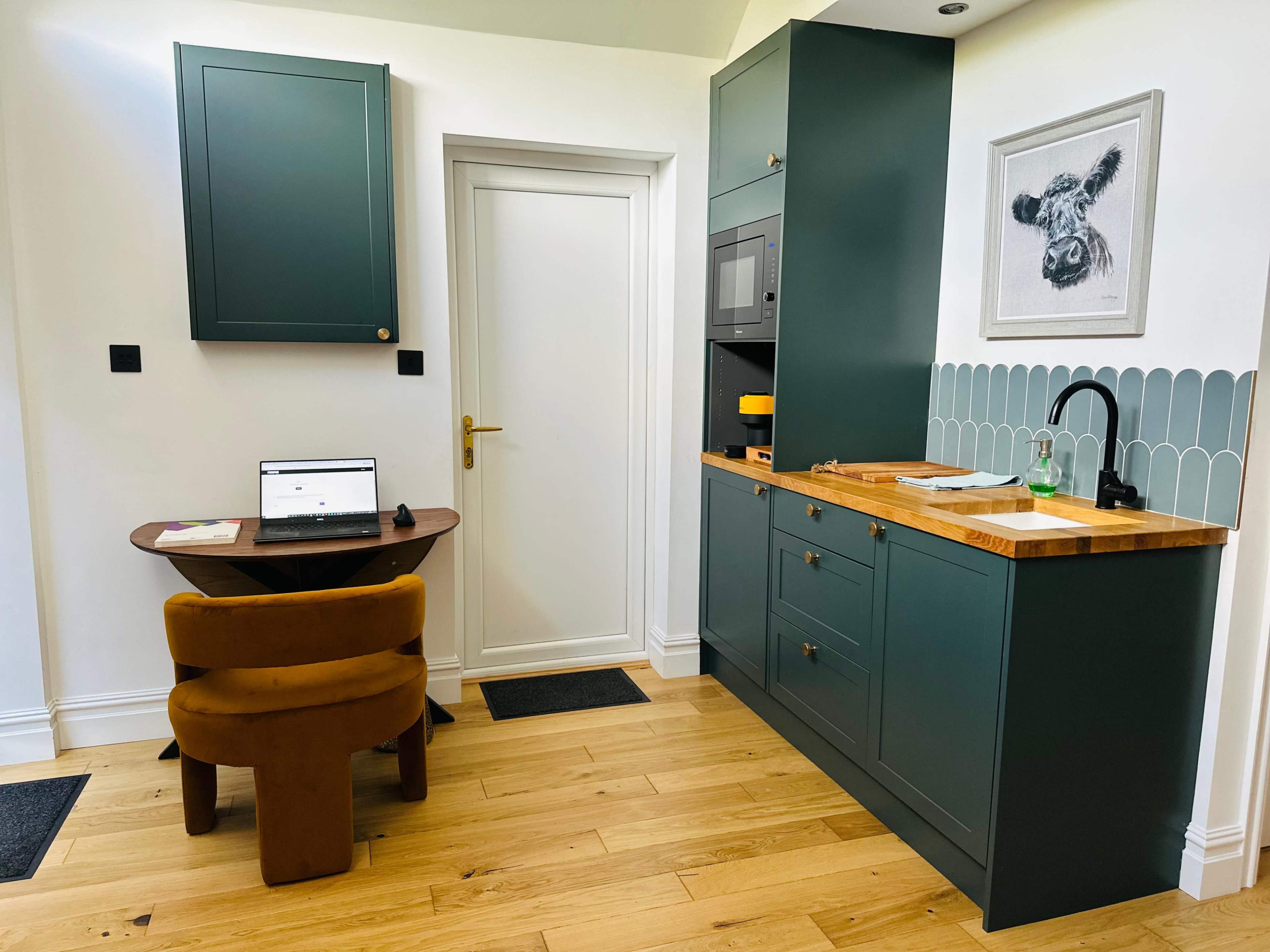 A modern kitchen area features a compact wooden table with a laptop, a dark green cabinet, a microwave, and a wall art of a cow.