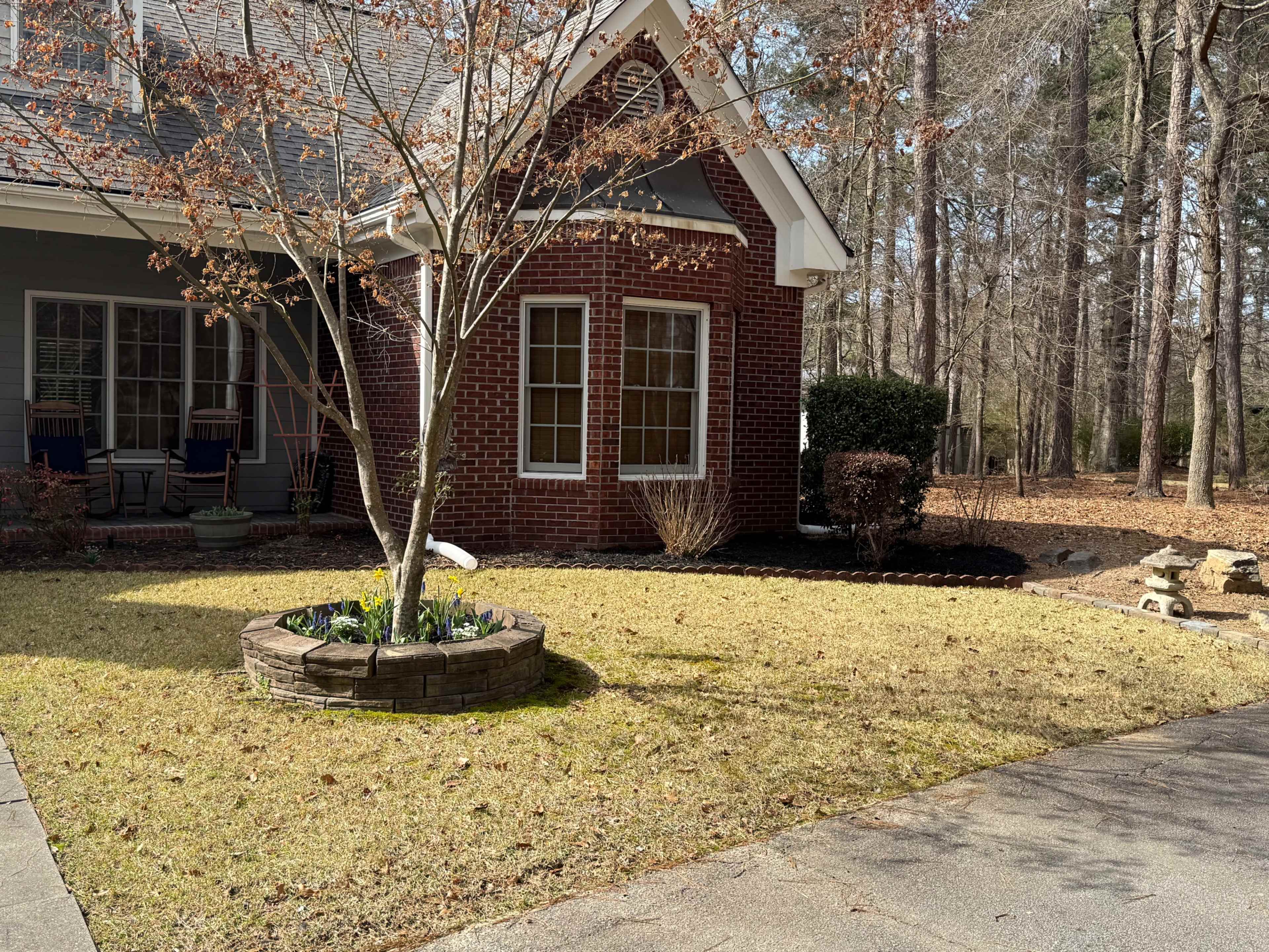 The image shows a brick house with a circular garden bed containing green plants, surrounded by a lawn and bordered by trees in a wooded area.
