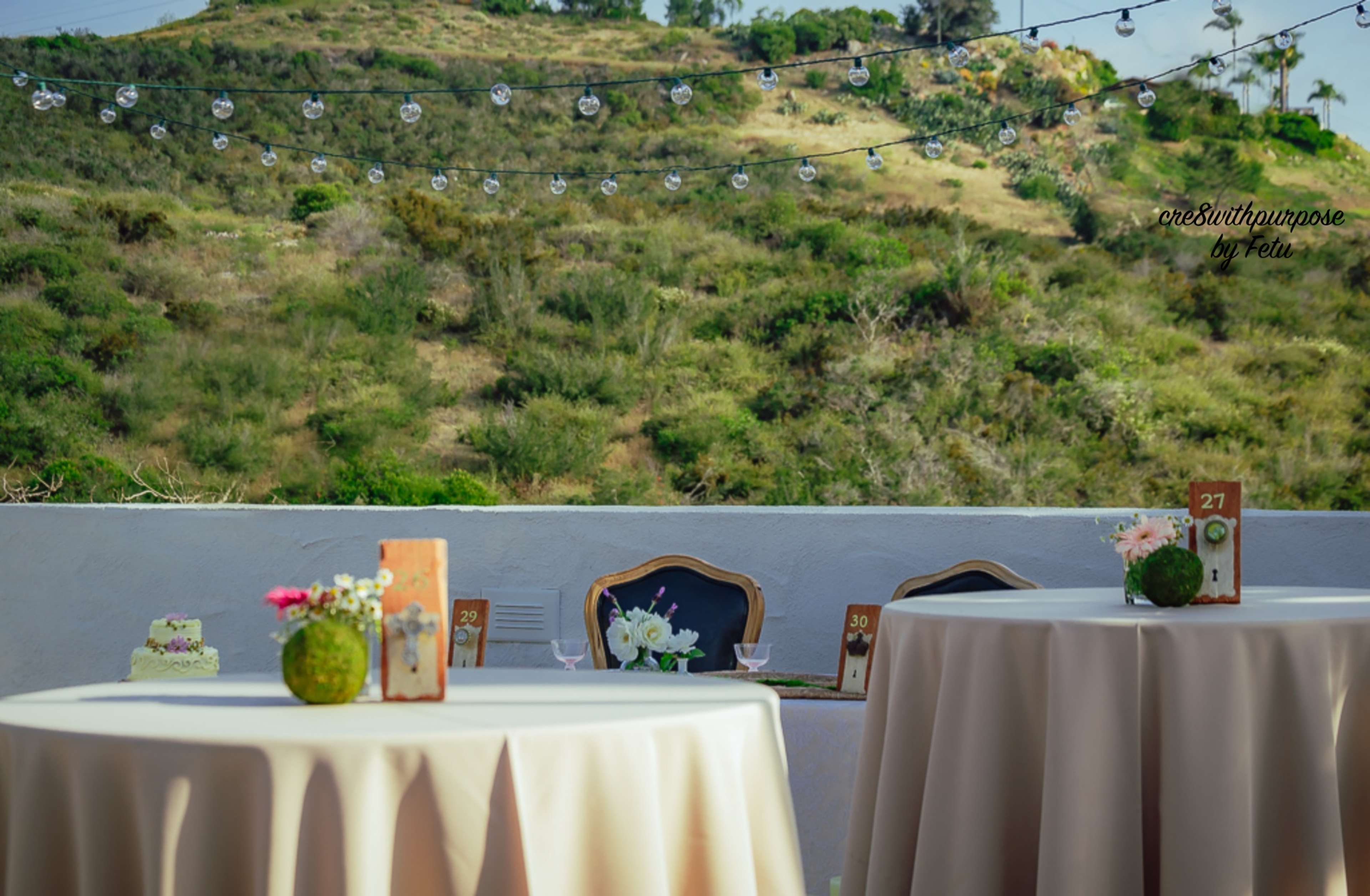 The scene features elegantly set round tables with light tablecloths, adorned with centerpieces, against a backdrop of a green hillside and string lights overhead.