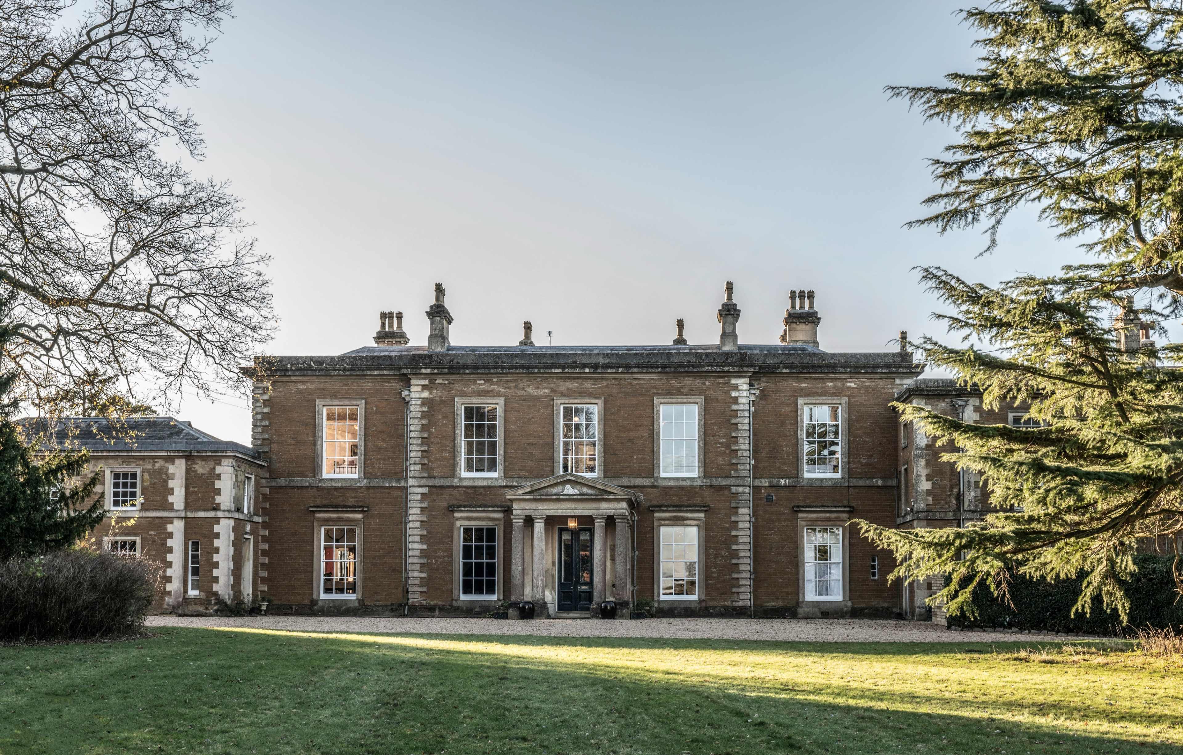 The image shows a large, two-story brick house with a symmetrical facade, featuring a central entrance and surrounded by a manicured lawn and trees.