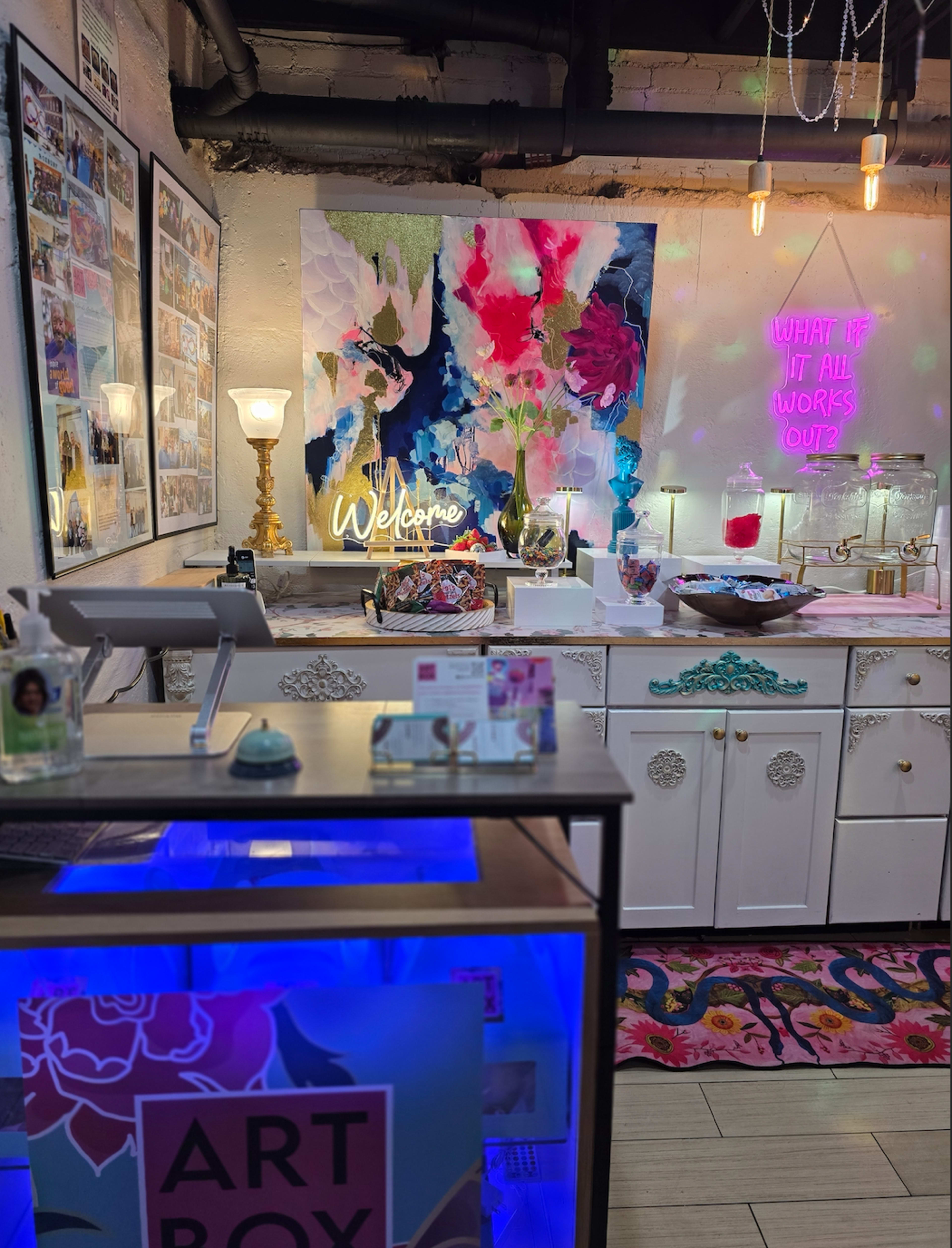 The image depicts a colorful art studio featuring a vibrant floral painting, a neon sign that reads "Welcome," and an assortment of art supplies and décor items arranged on a counter.