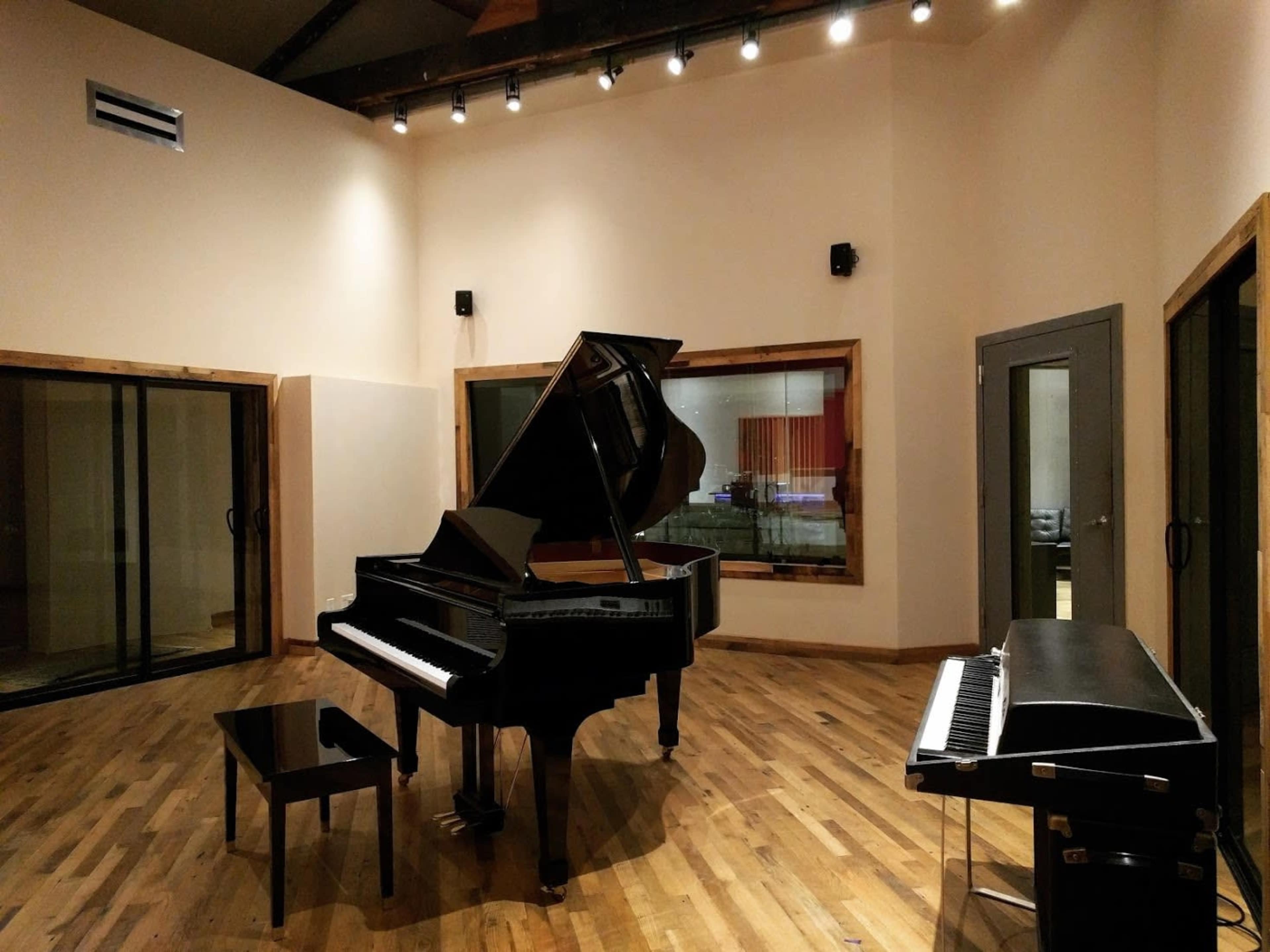 A grand piano and a small black upright piano are positioned in a spacious recording studio with wooden floors and large glass doors.